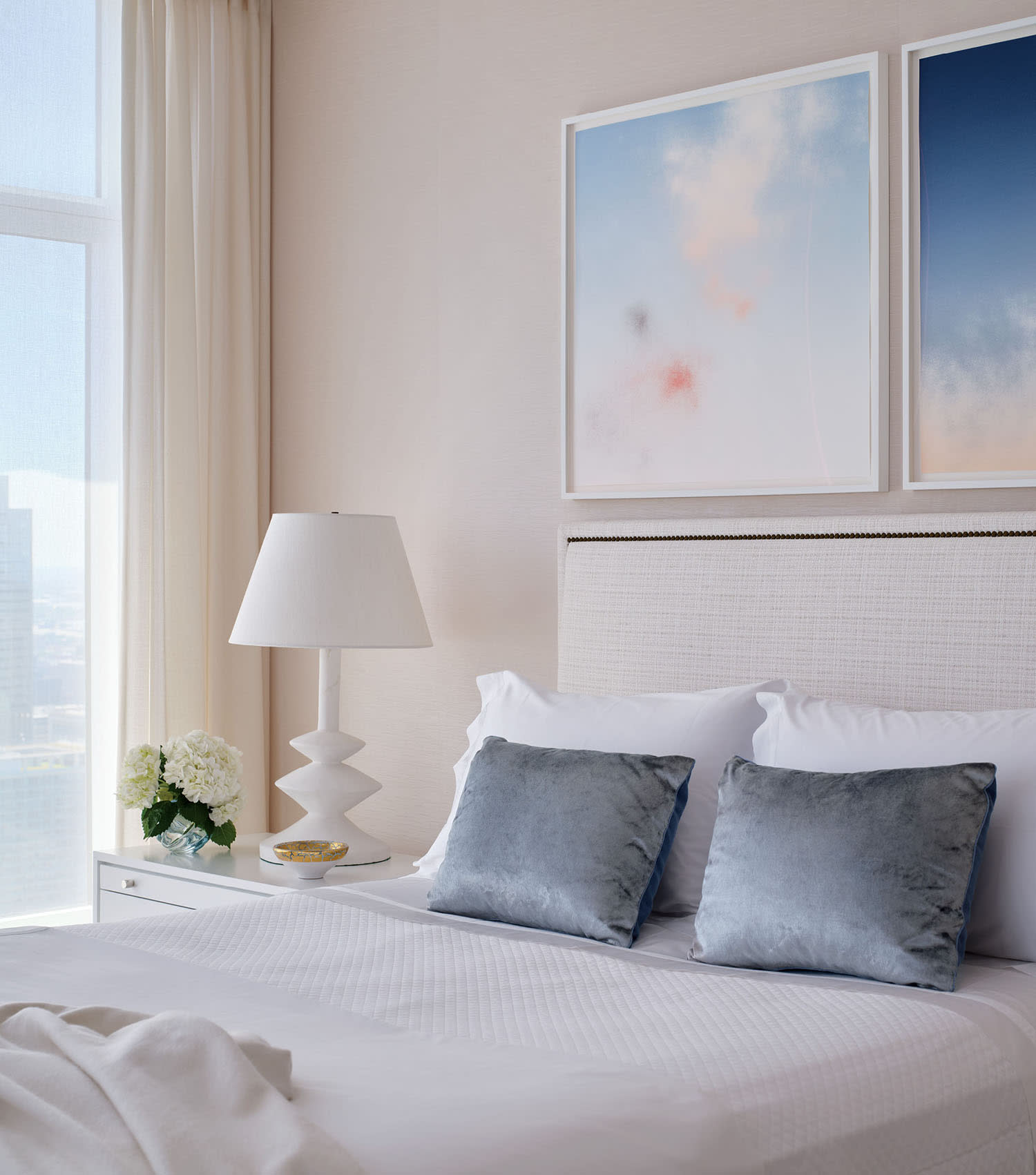 A bed made up with white linens and gray accent pillows, with two framed artworks mounted above.