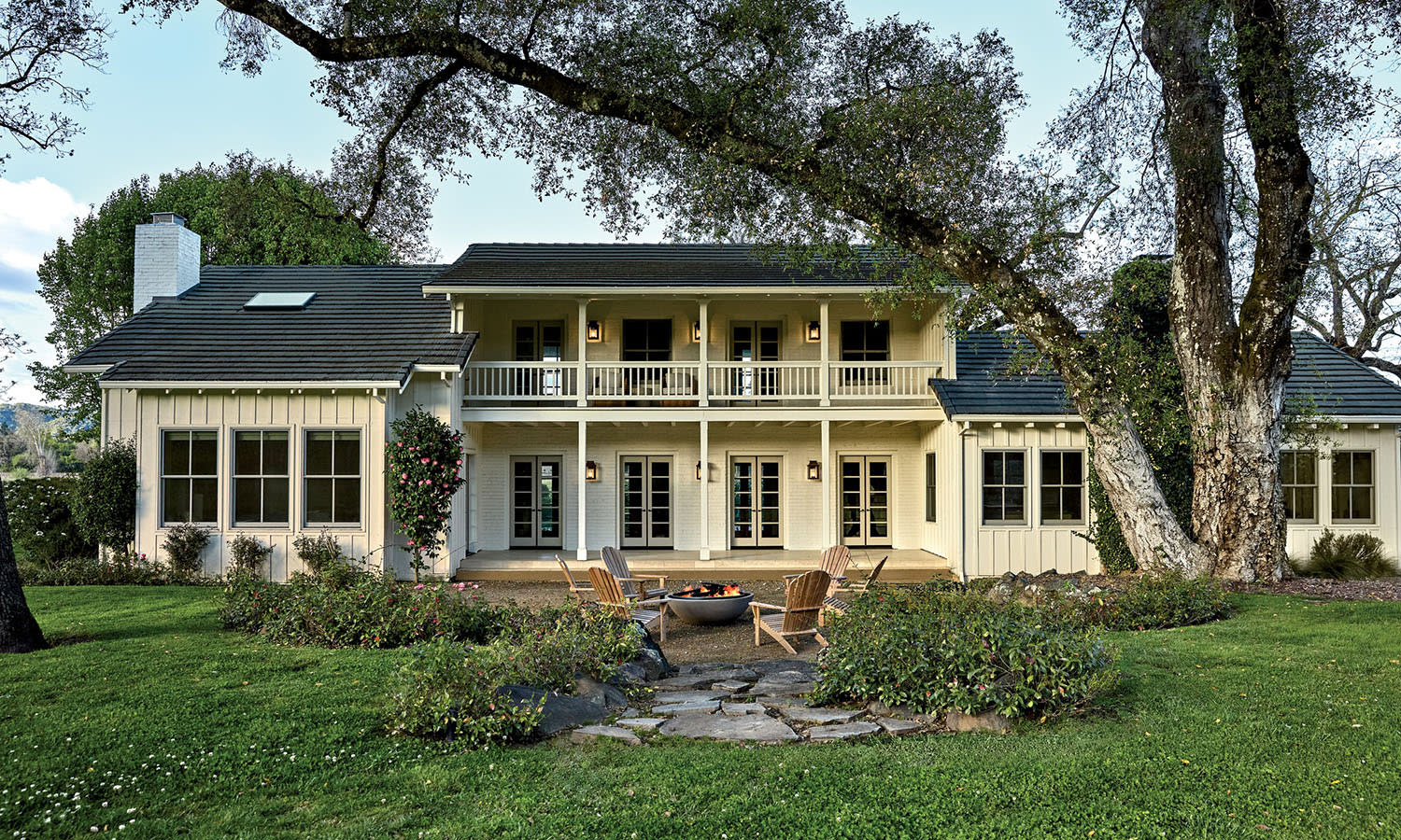 Two-story farmhouse with a front porch and balcony, surrounded by lush greenery. Cozy chairs encircle a fire pit on a stone patio, inviting relaxation.