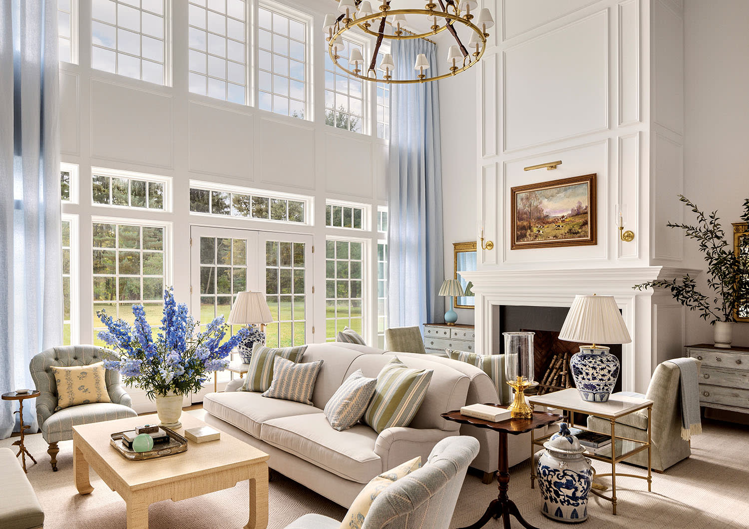 Elegant living room with high ceilings, large windows, and soft blue curtains. Features a grand chandelier, cozy white sofa, and floral accents.