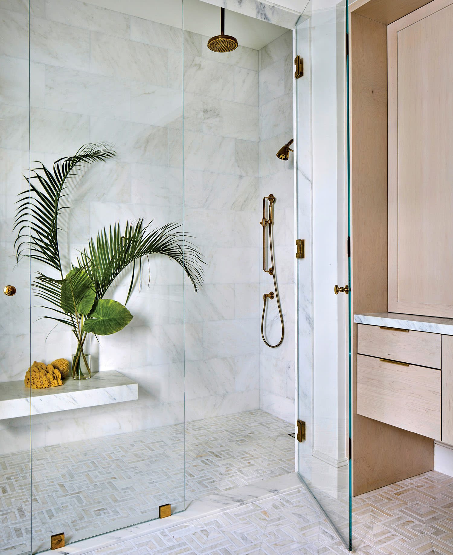 Luxurious bathroom with a marble-tiled shower, gold fixtures, and glass doors. A large green plant adds a touch of nature, enhancing the serene ambiance.