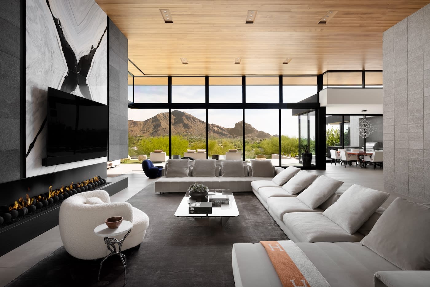 Modern living room with a large, angular beige sofa, sleek fireplace, and floor-to-ceiling windows revealing a mountain view. Neutral tones create a serene atmosphere.