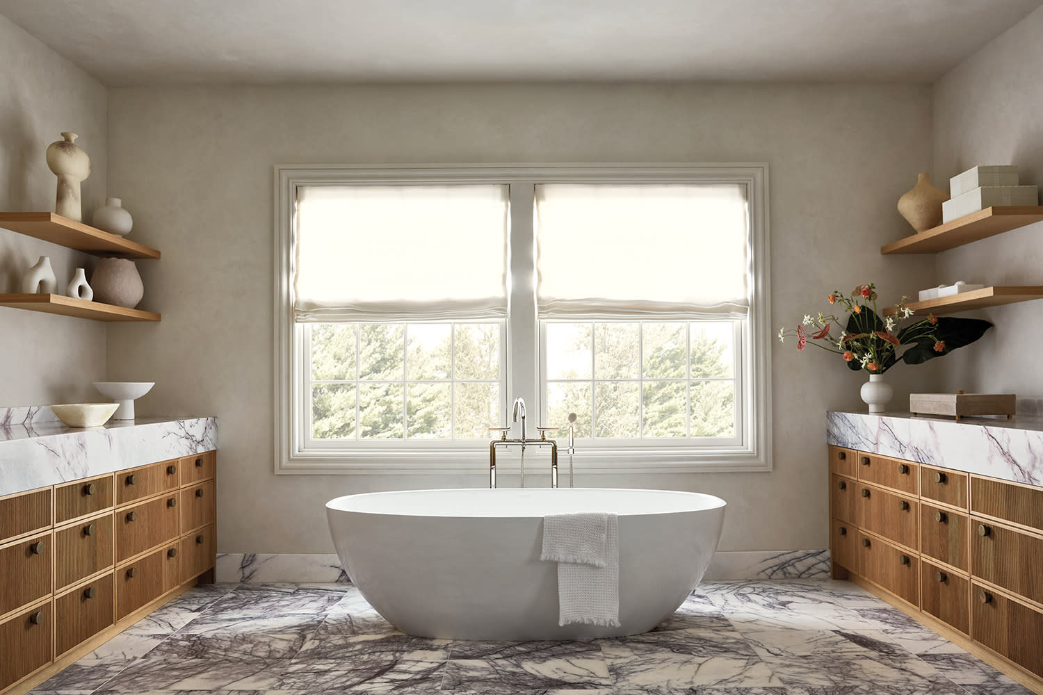 Freestanding tub in a bright bathroom with marble floor, twin wood vanities, and window.