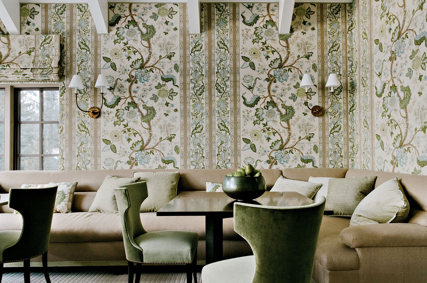 Wallpaper with a pattern of green leaves covers a room with a couch, tables and chairs.