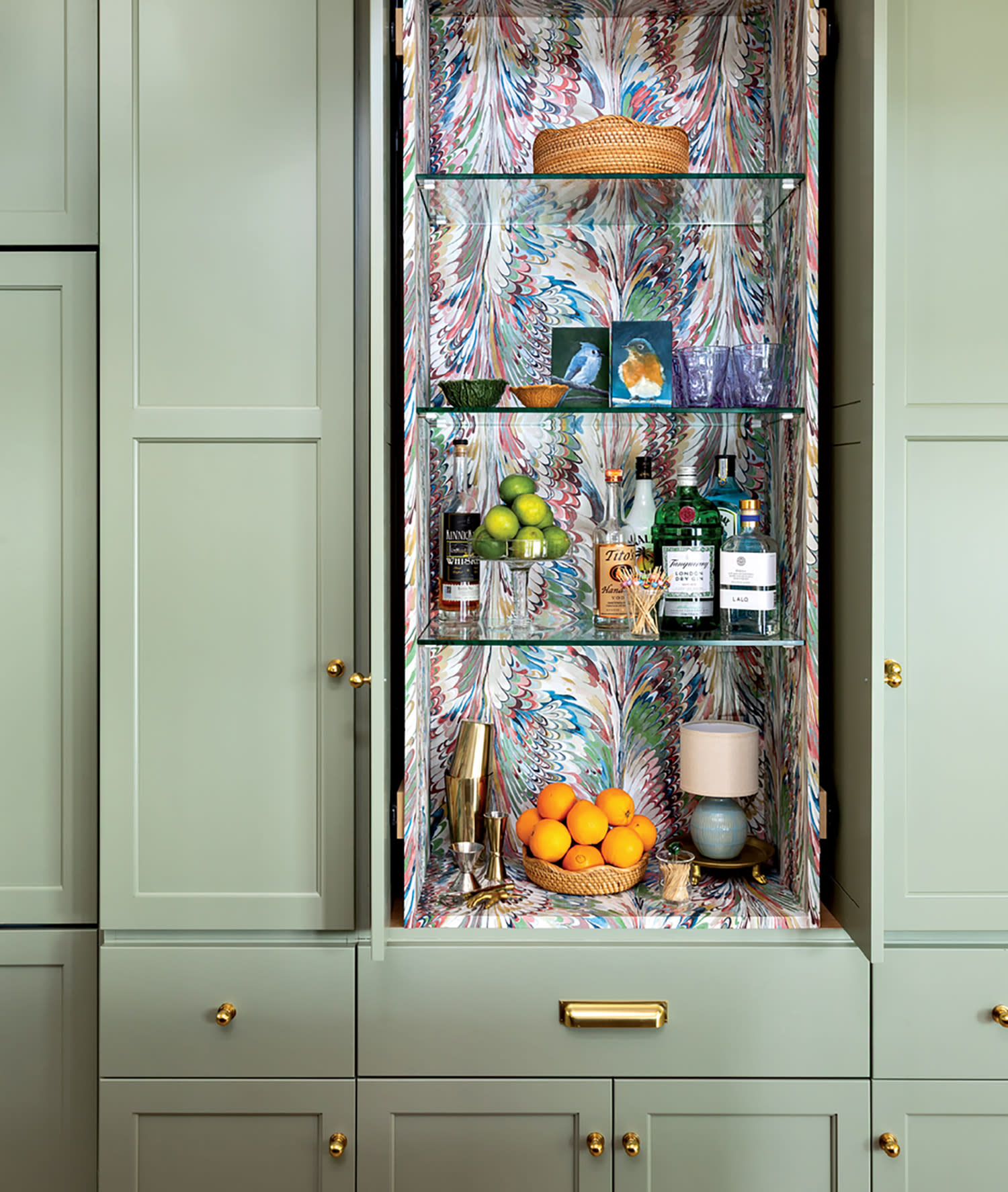 Elegant bar cabinet with open green doors revealing a colorful, marbled interior. Shelves hold fruits, bottles, glassware, and a small lamp, creating a chic and vibrant display.