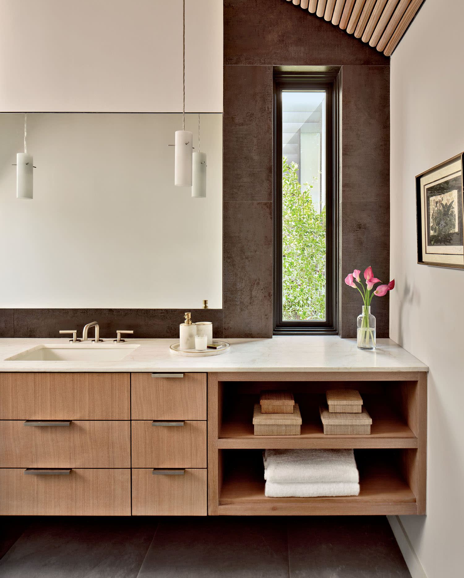 Modern bathroom with a wooden vanity, large mirror, and pendant lights. A vase with pink flowers and two towels are neatly placed on the counter.