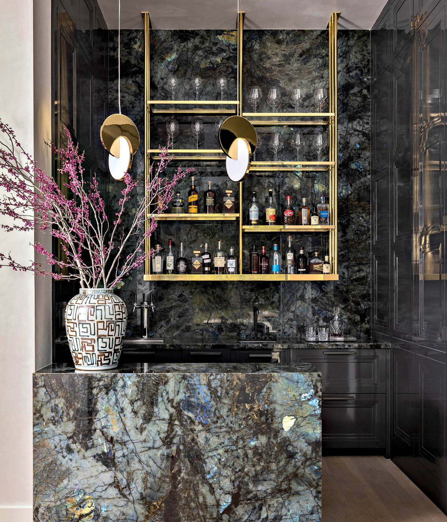 A cocktail bar with stocked wall shelves, gray cabinets and a granite bar decorated with a vase. 