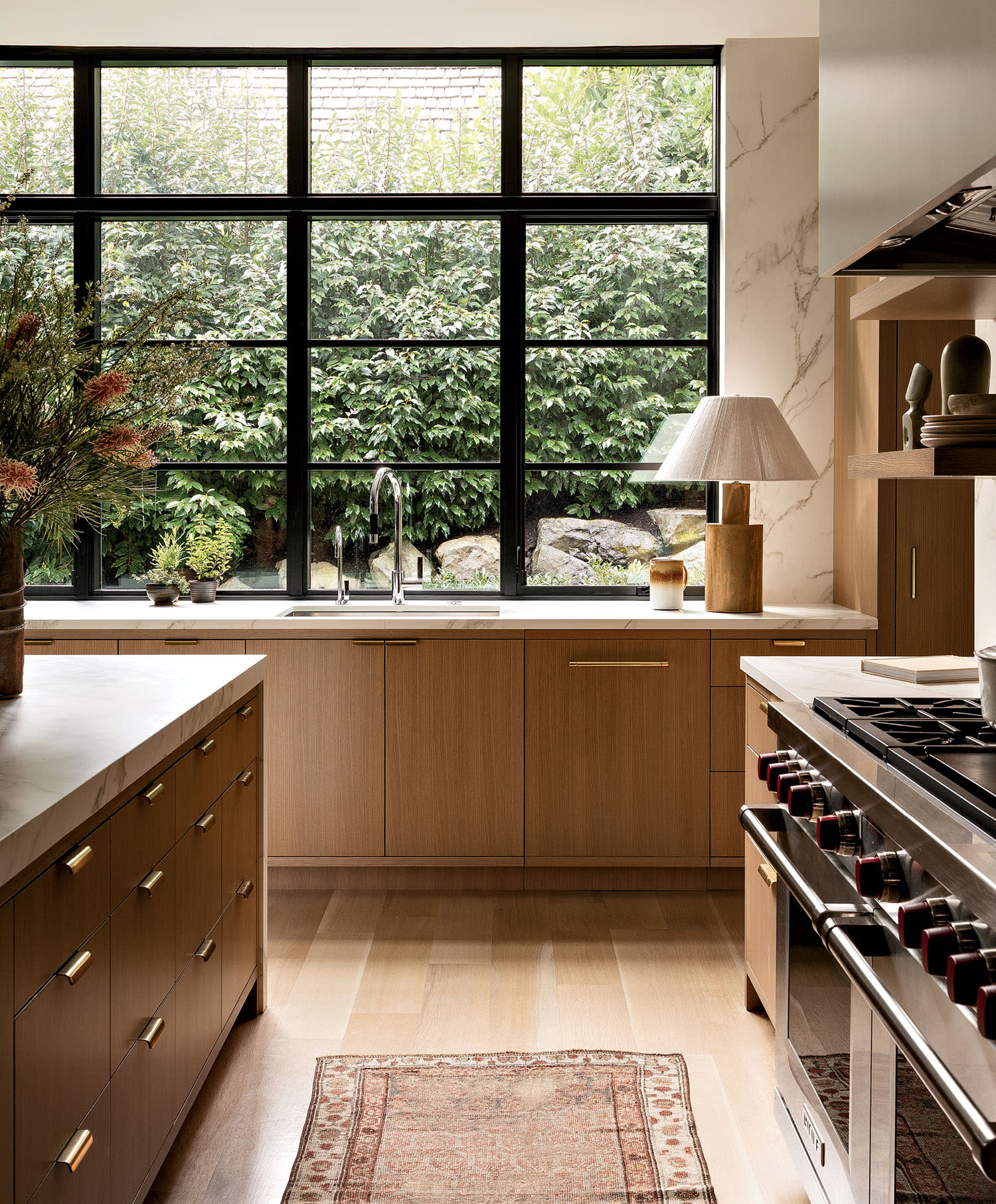 Warm, modern kitchen with wood cabinets, a large window revealing lush greenery, and a central island. A cozy rug and sleek appliances add style.