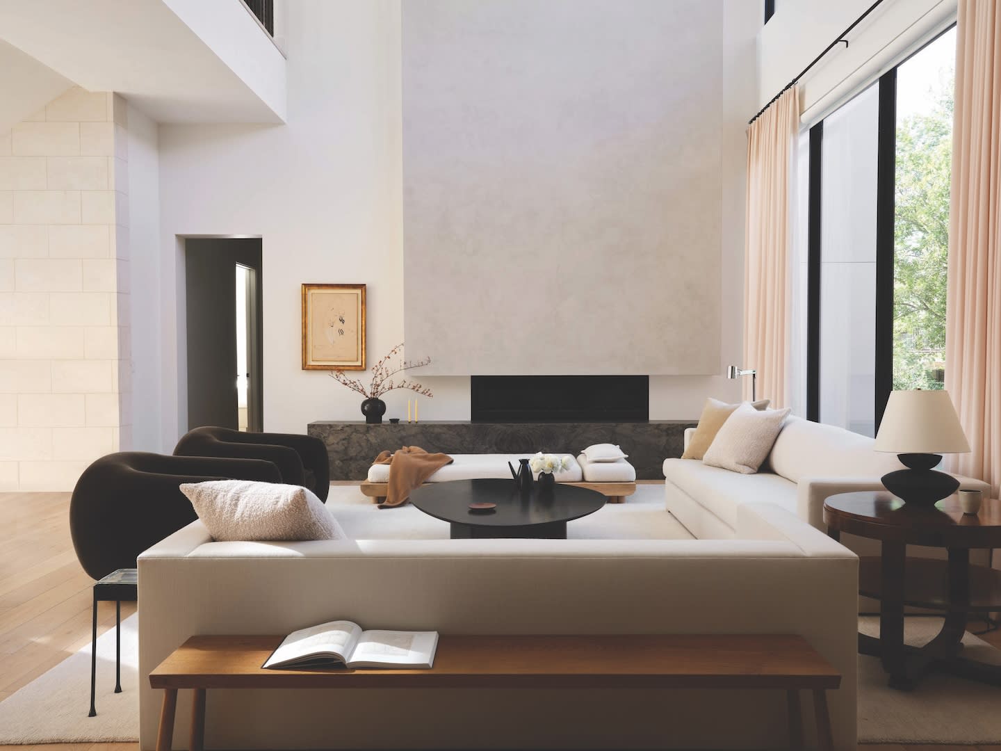 Elegant living room with a high ceiling, featuring white sofas, a black round coffee table, modern decor, soft lighting, and large windows with beige curtains.