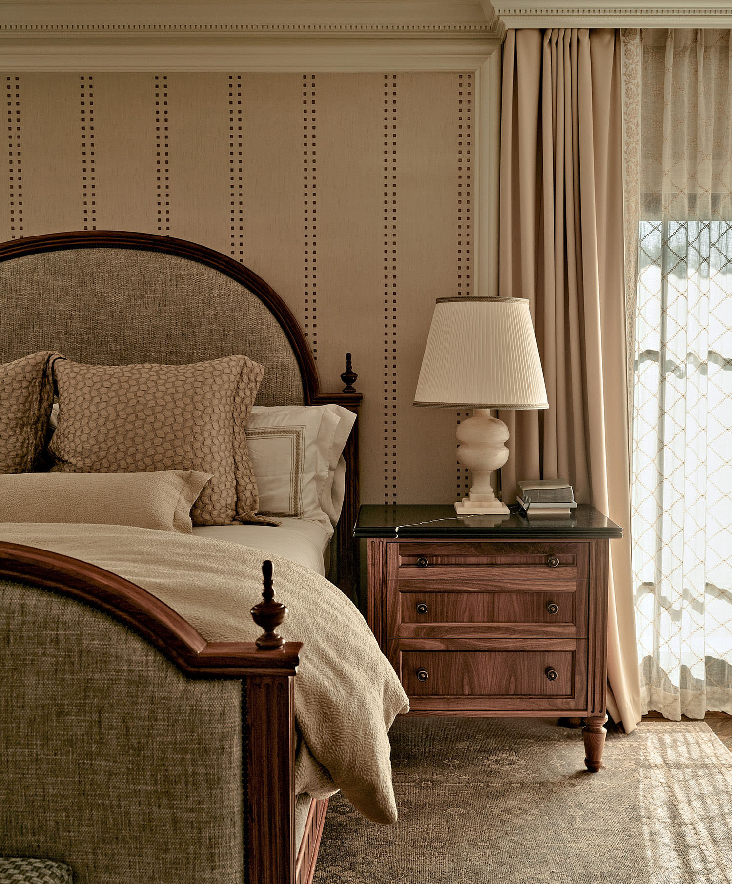 Cozy bedroom with a wooden bed featuring beige sheets and pillows. A matching nightstand holds a lamp and books. Soft light filters through sheer curtains.