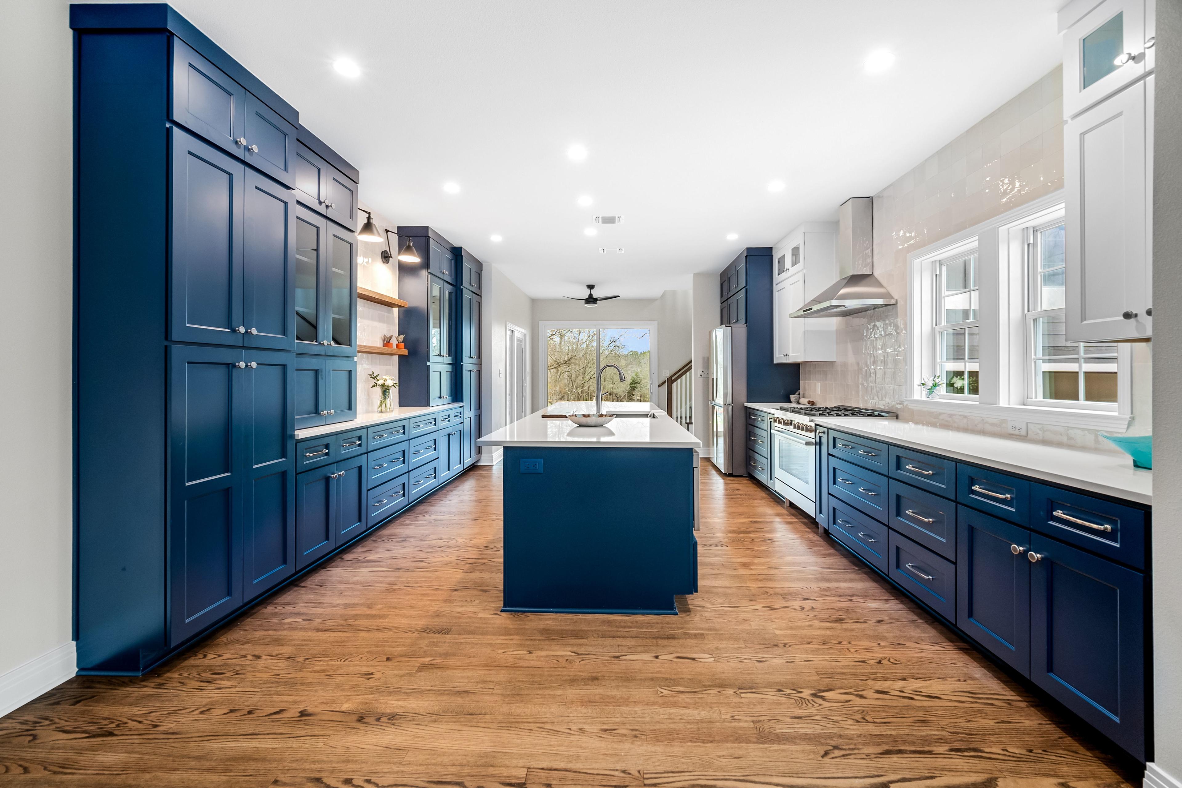 This vibrant kitchen features bold navy cabinetry, sleek white countertops, and stainless-steel appliances. A central island anchors the space, while natural wood flooring and open shelving create a perfect balance of modern elegance and warmth.