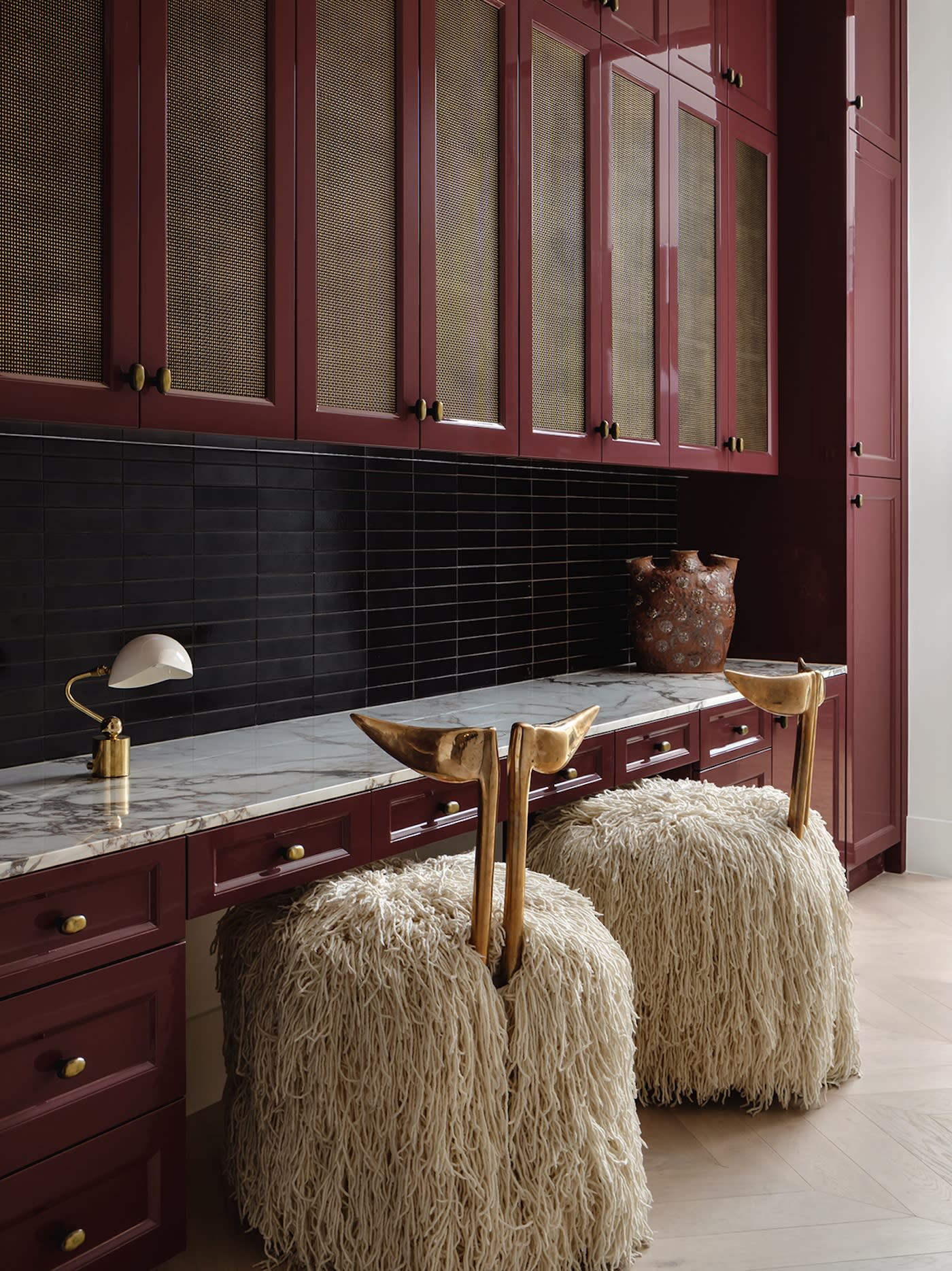 Elegant kitchen with dark red cabinets and woven doors, marble countertop, black tile backsplash, and two textured, shaggy stools with brass accents.