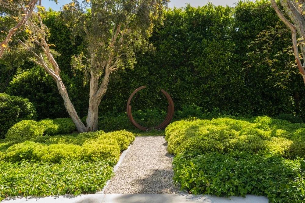 A tranquil garden scene with a gravel path leading to a circular metal sculpture, surrounded by lush greenery and trees, evoking a serene atmosphere.