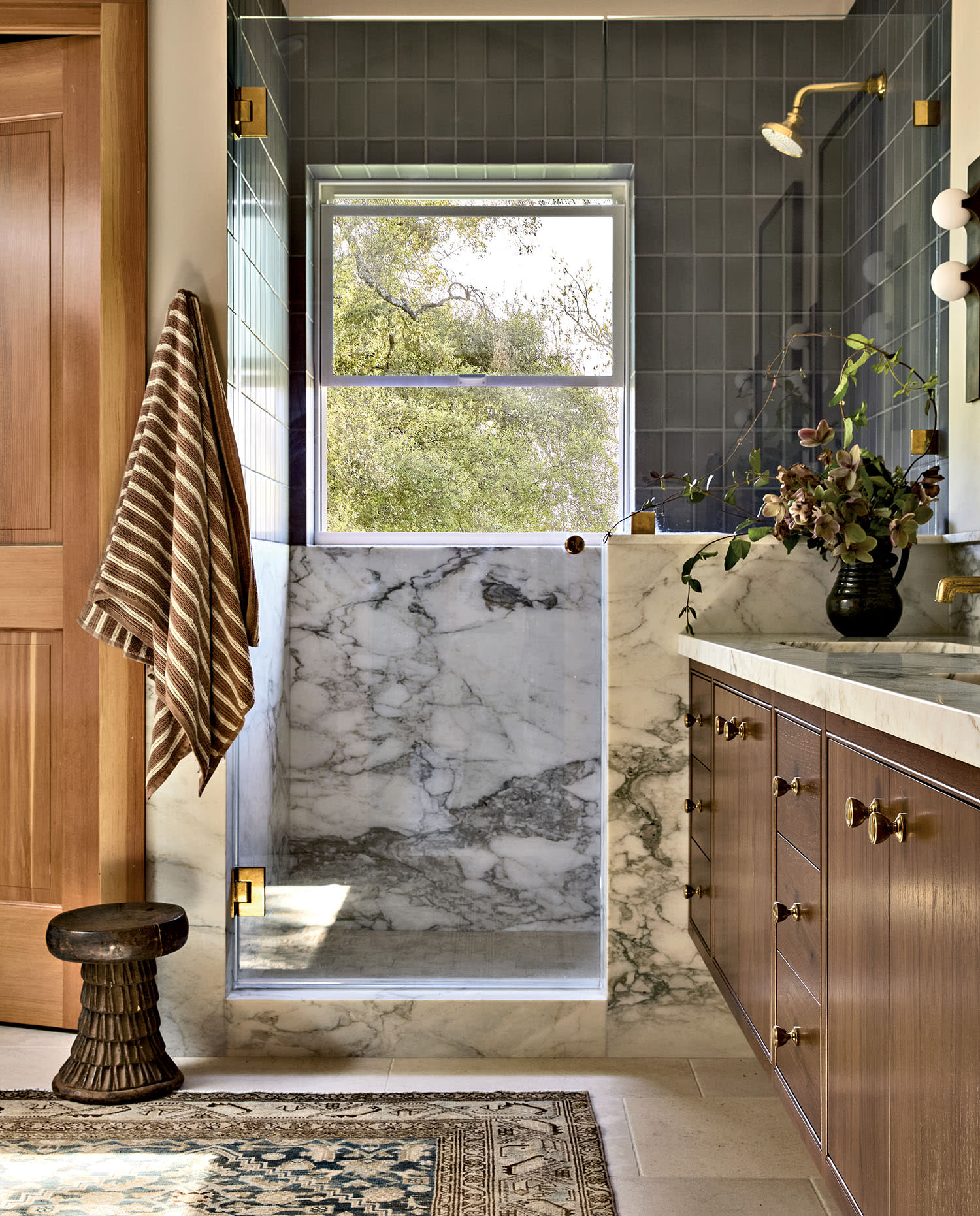 Luxurious bathroom featuring a marble shower and countertop, wood cabinets, and a striped towel. A large window offers a view of green foliage.