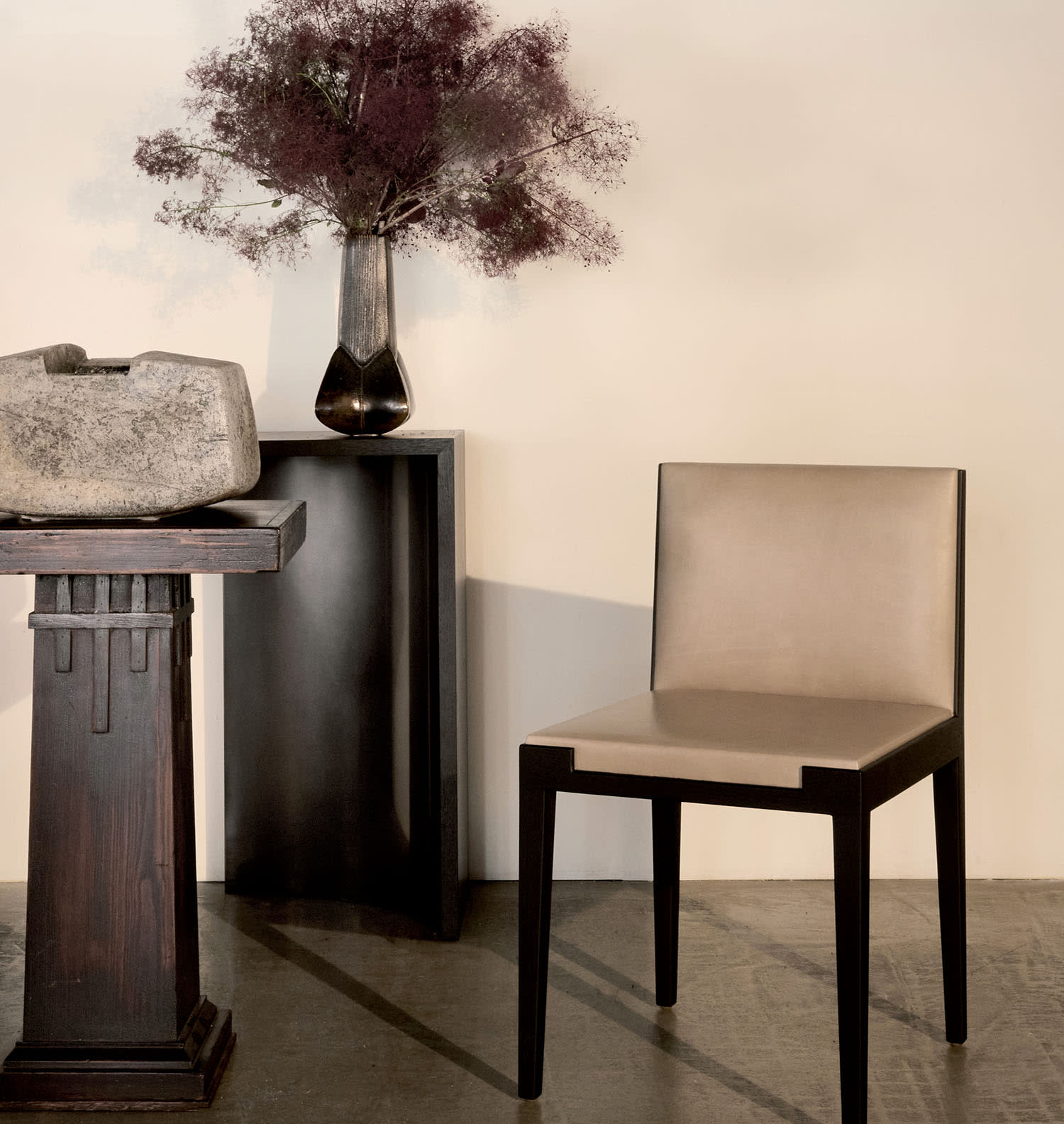A chair with dark legs and light upholstery sits on a wood floor near a sculpture and a plant.