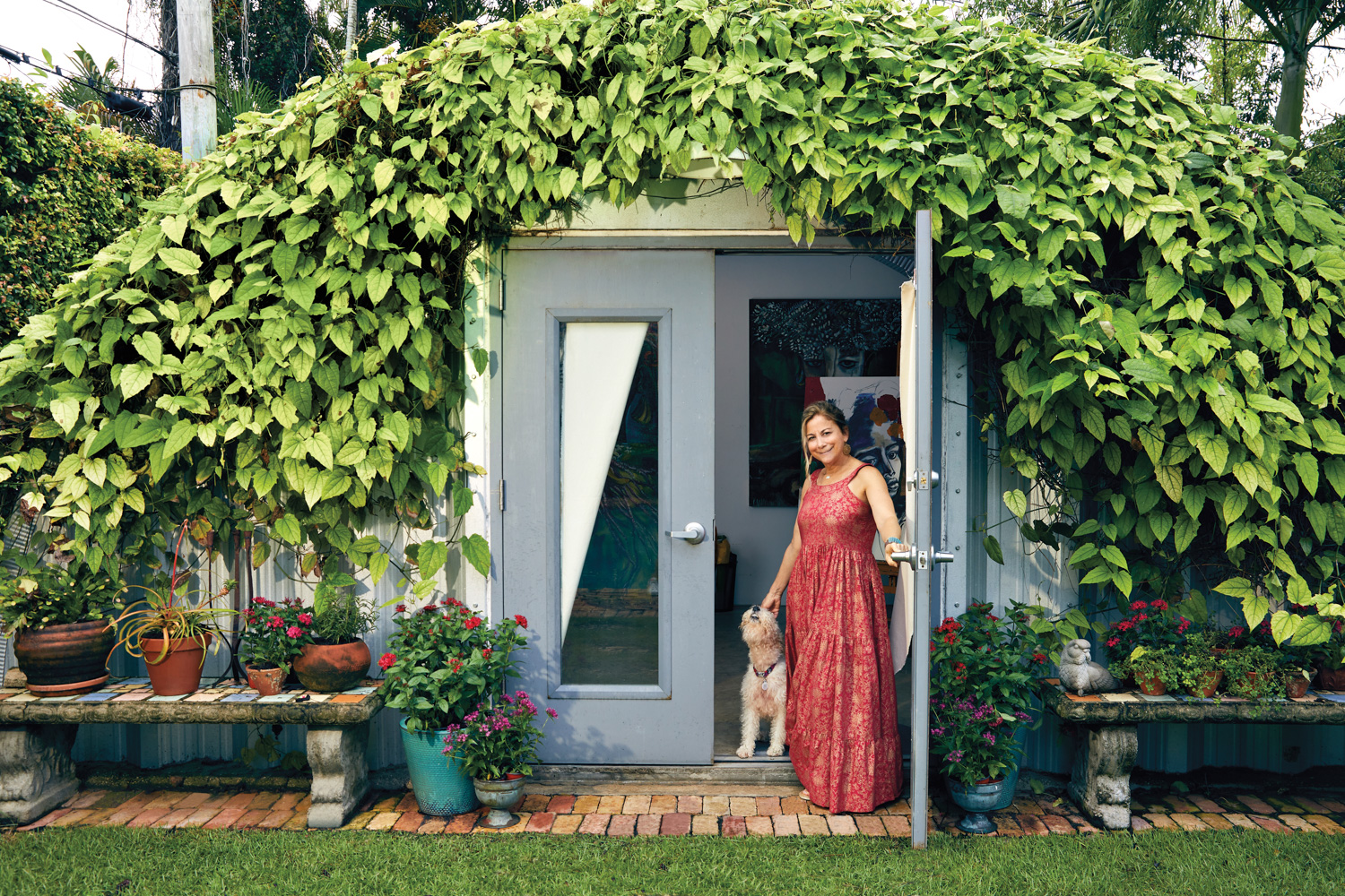 artist ana trelles portuondo with her wire fox terrior outside her art studio covered in a flowering vine