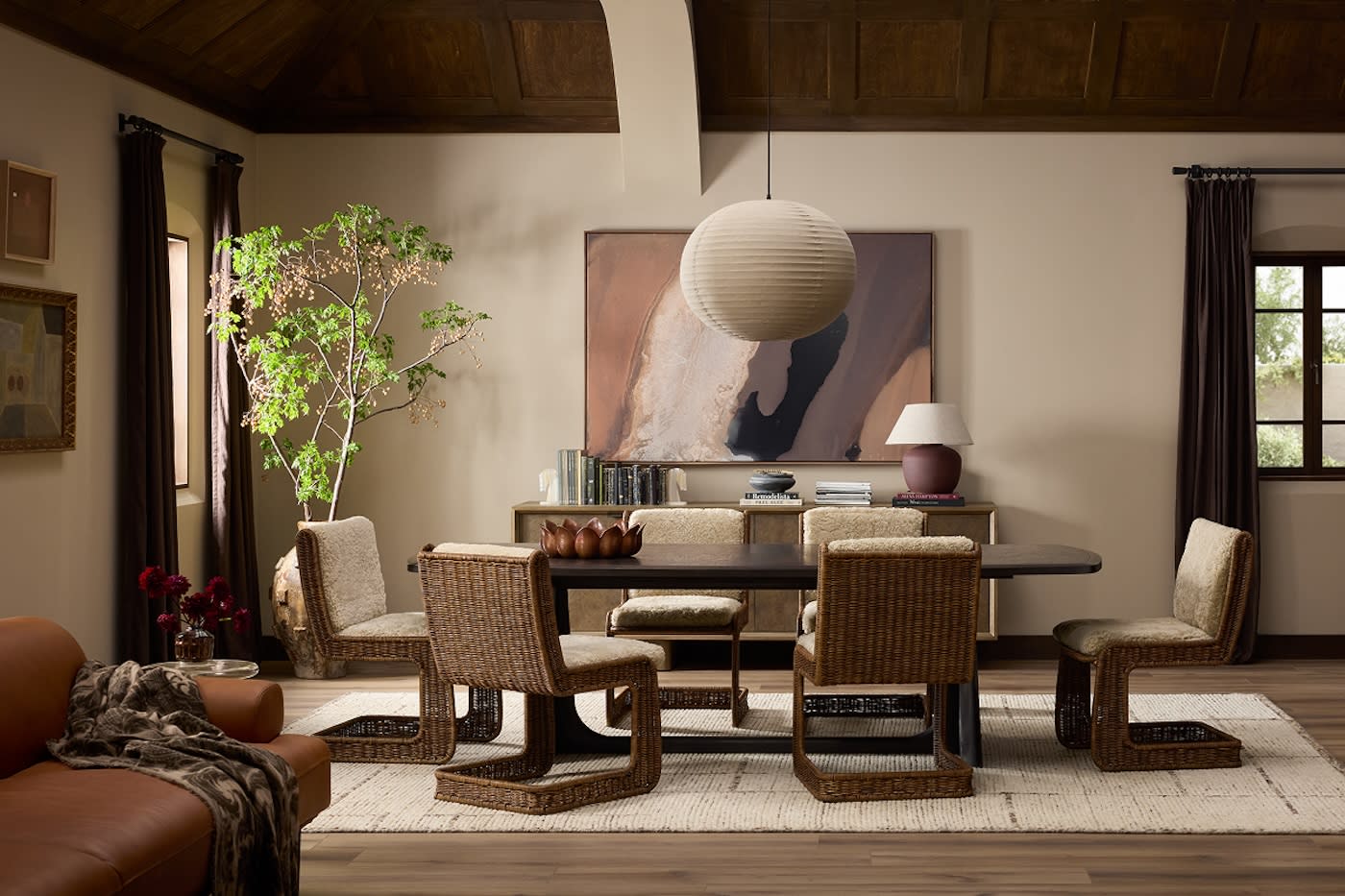 Warm, cozy dining room with wicker chairs around a dark table, beige walls, large abstract painting, round pendant light, and lush plant.