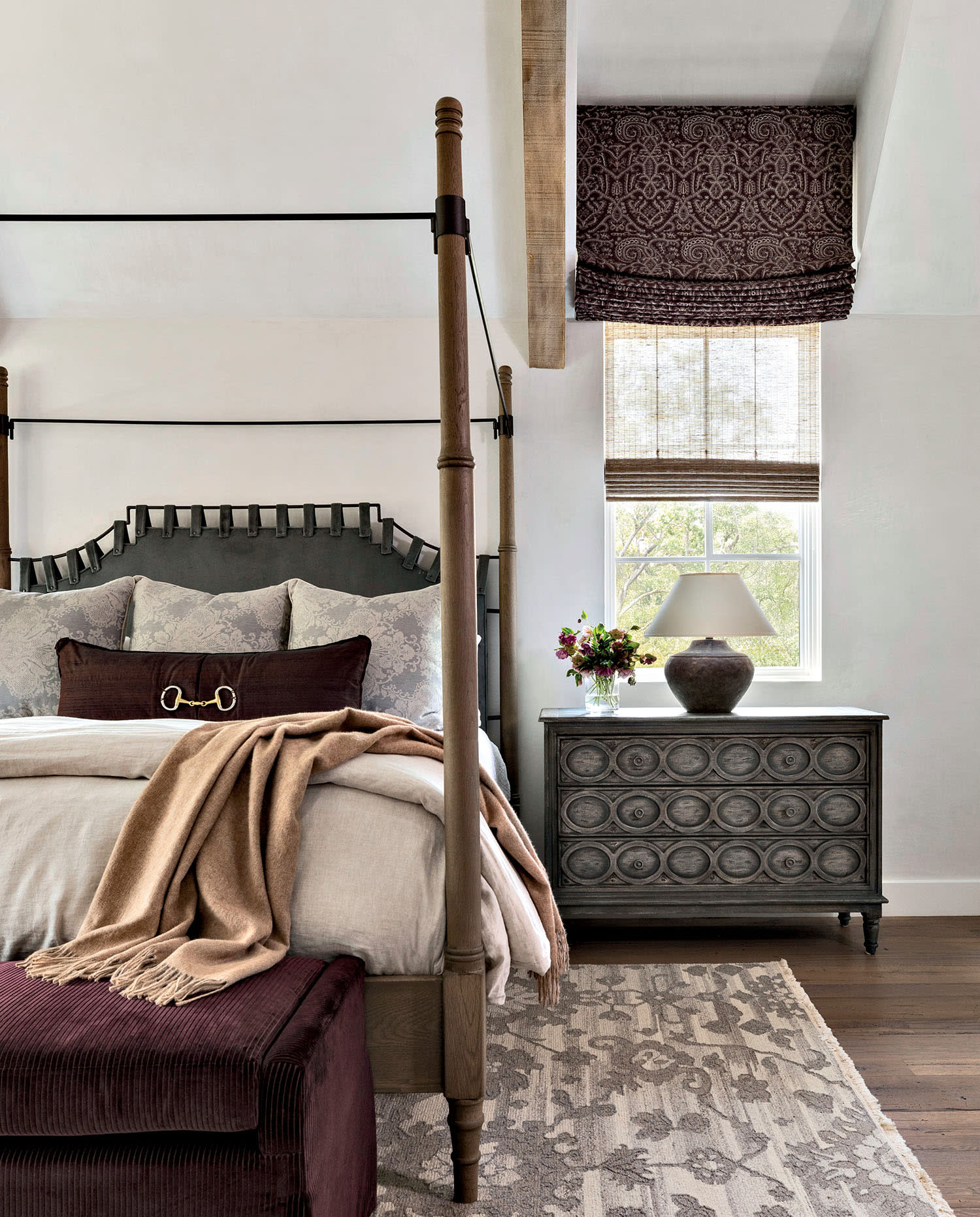 Elegant bedroom with a wooden four-poster bed, beige bedding, and a brown throw. A patterned rug and carved dresser add a cozy, sophisticated touch.