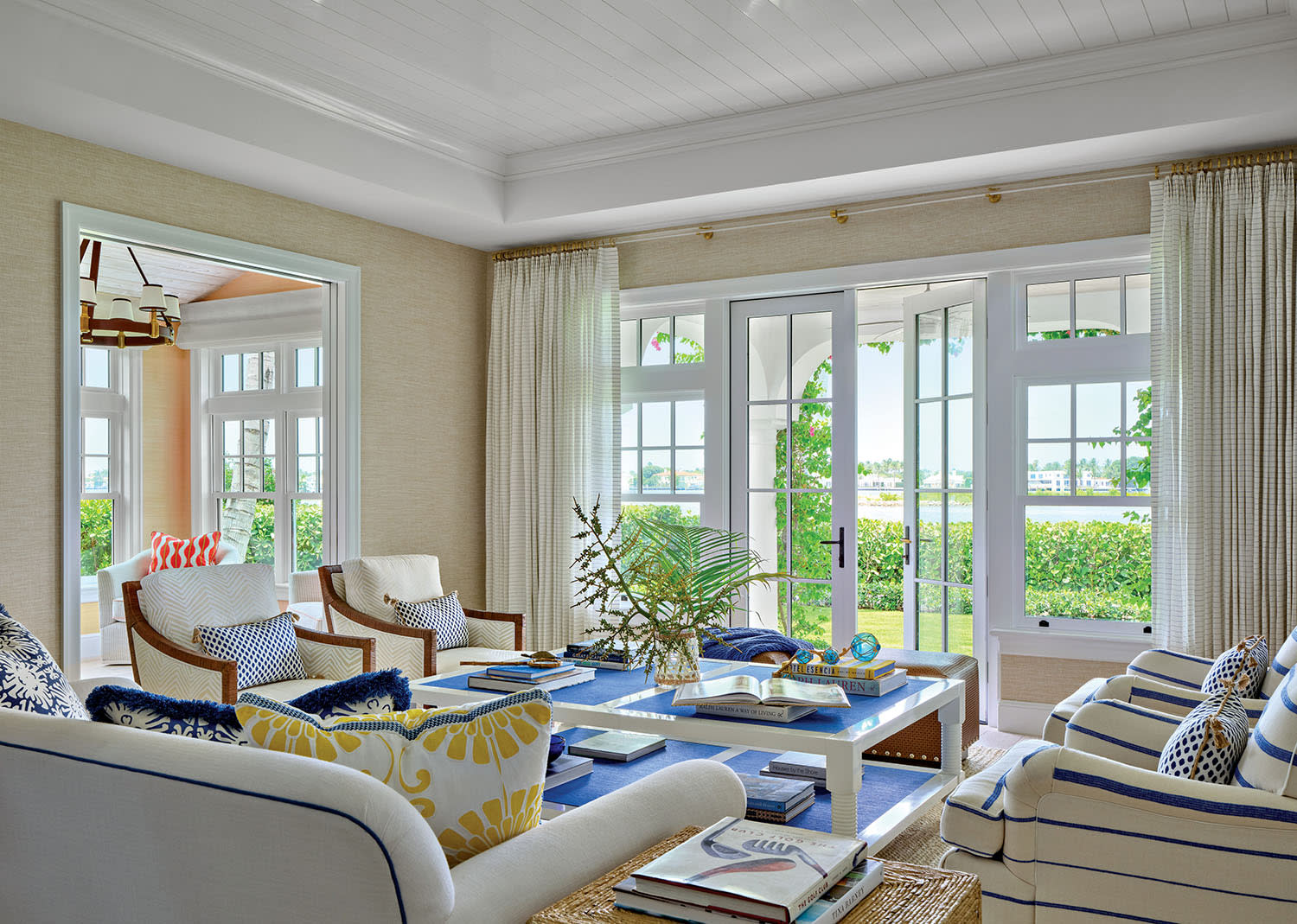Bright, coastal living room with large windows, light furniture, and nautical accents. A blue coffee table is adorned with books, creating a cozy, inviting feel.