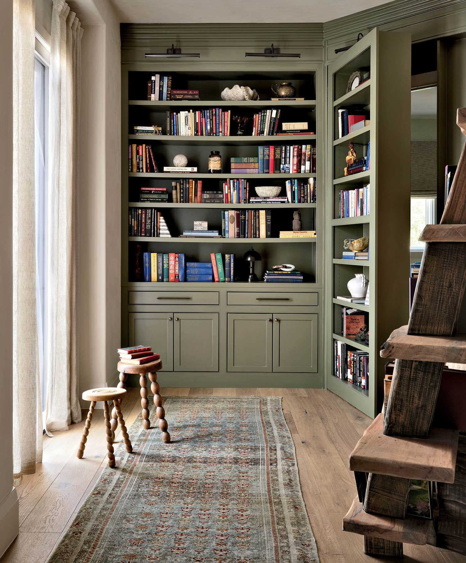 Cozy room with a tall, sage green bookshelf filled with books and decor. A wooden ladder leans against the shelf. Warm light streams through curtains, illuminating the patterned rug and wooden stools.