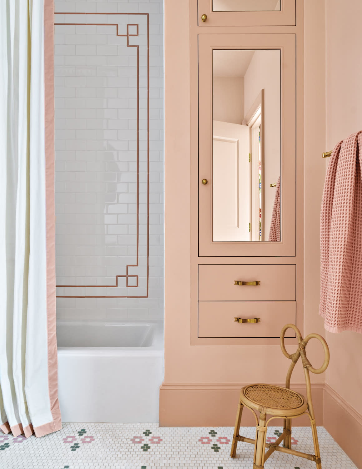 Soft pink bathroom with elegant design. Features a white tub, geometric tile accents, a wicker chair, and a mirrored cabinet, creating a cozy atmosphere.