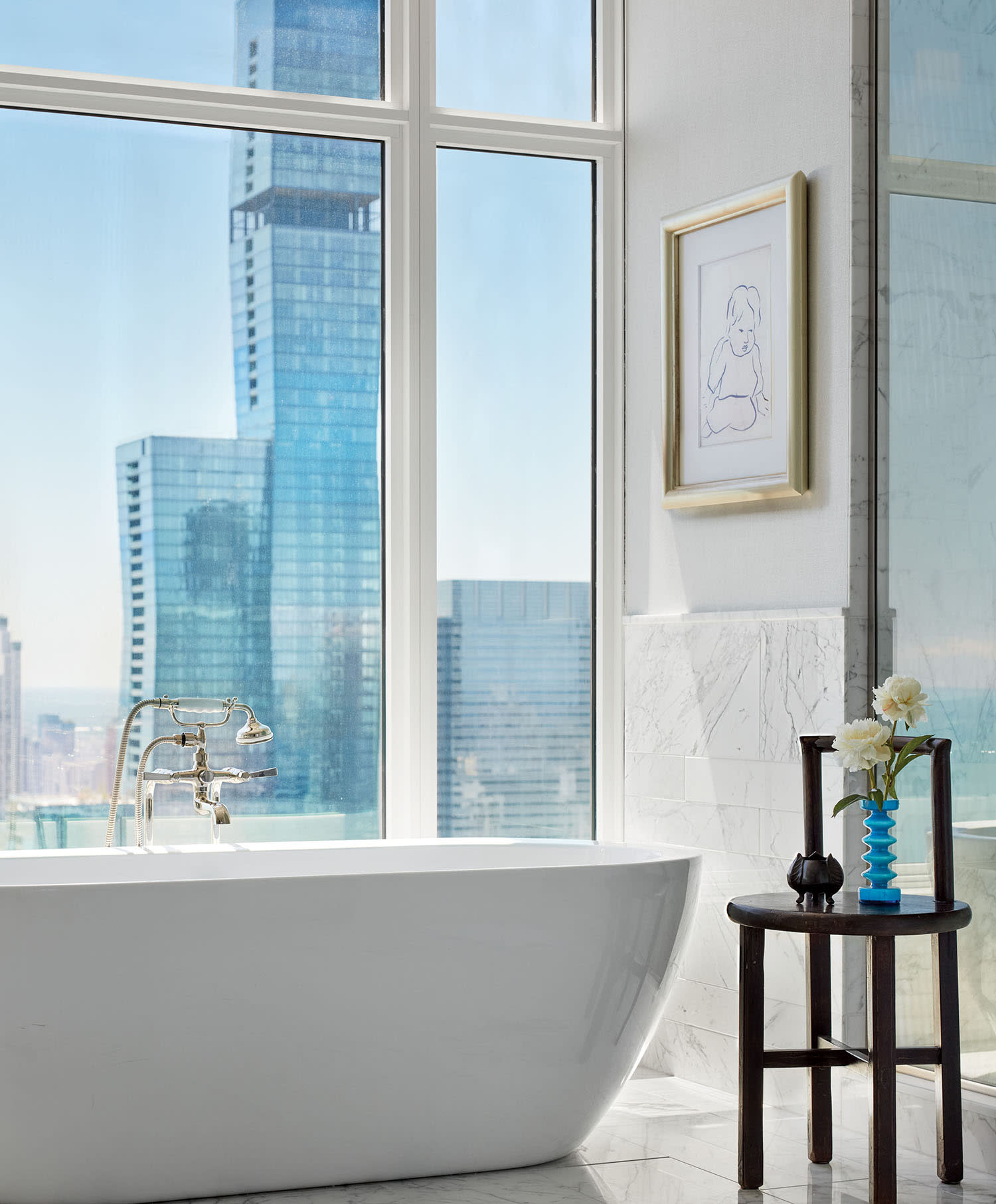 A windowed bathroom with skyline views, a white bathtub and wood accent table set with a small vase.