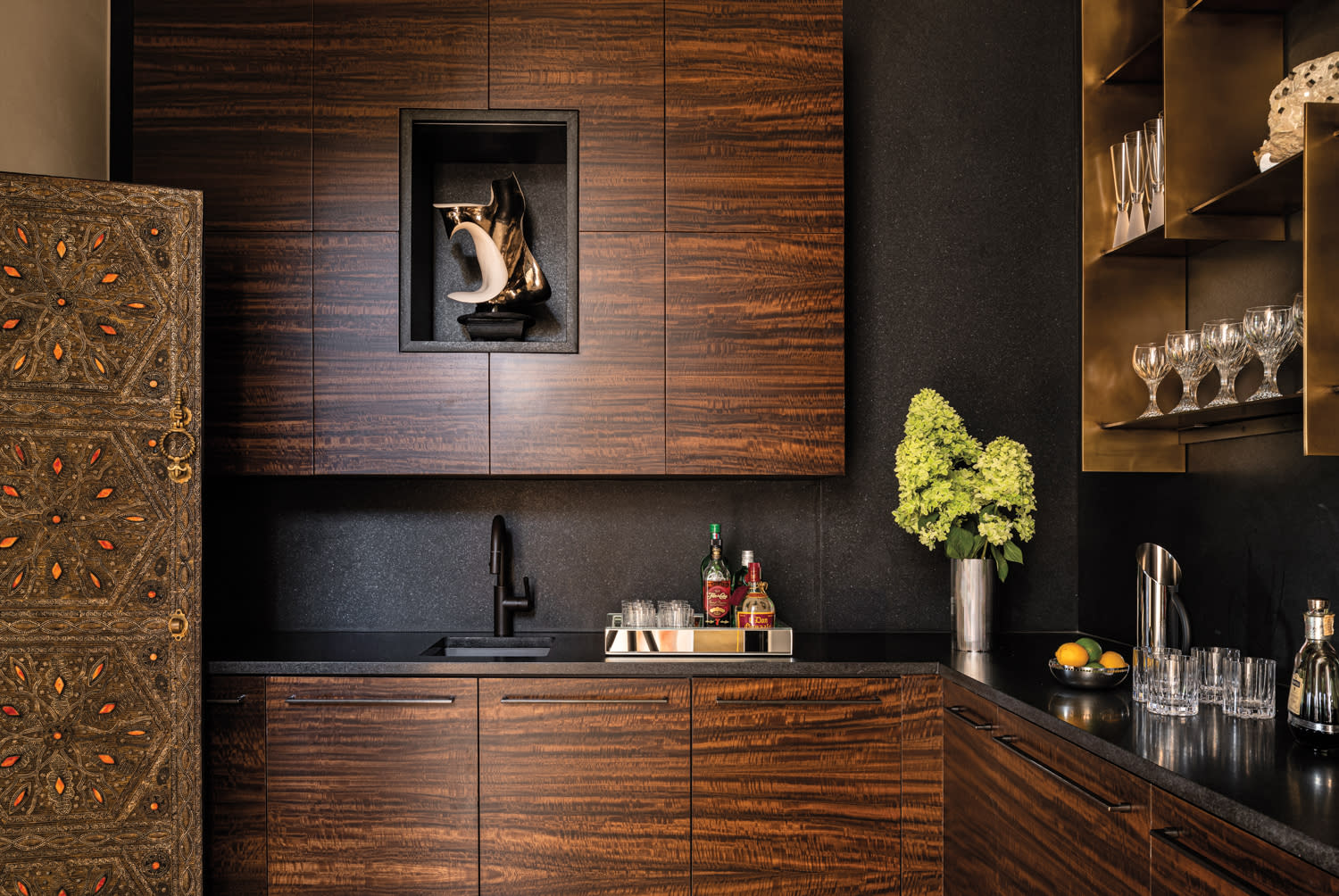 A modern kitchen with dark wood cabinets, a black countertop, and bronze open shelving. A vase with green flowers and a tray with liquor and glasses are on the counter.