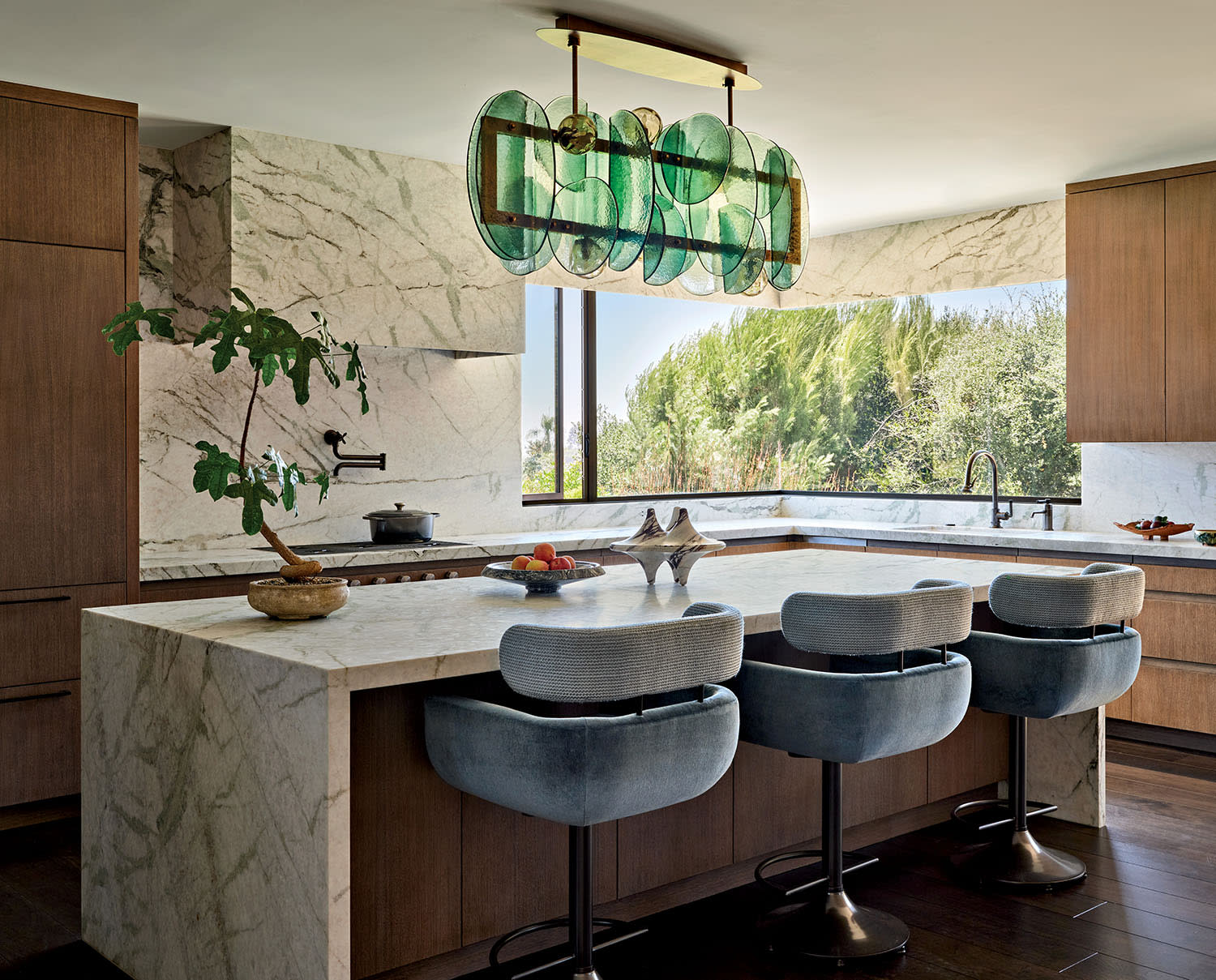 Modern kitchen with a marble island, sleek wooden cabinetry, and blue velvet chairs. A unique green glass chandelier hangs above.