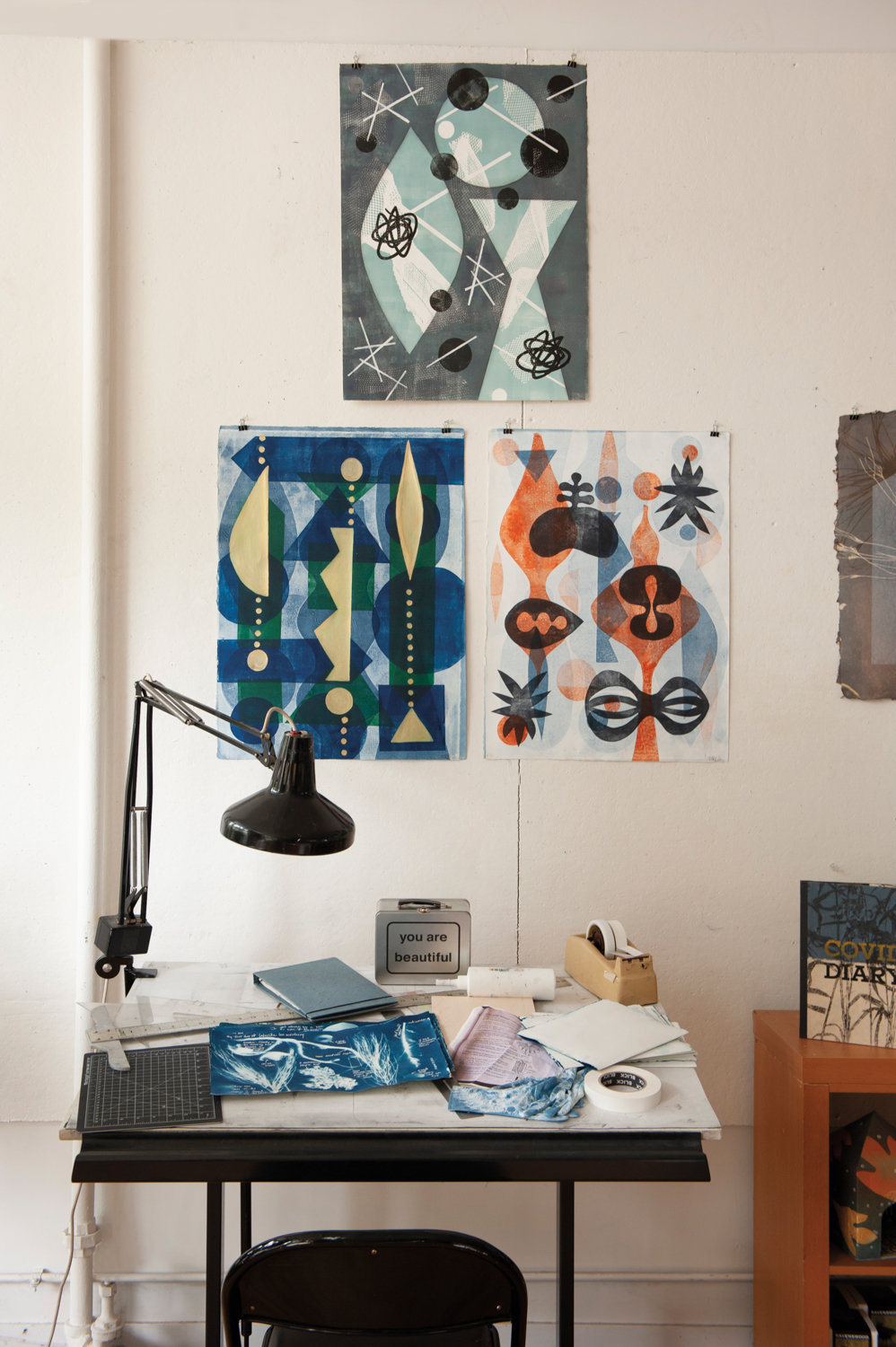 Three monoprints hang on a wall above a small desk.