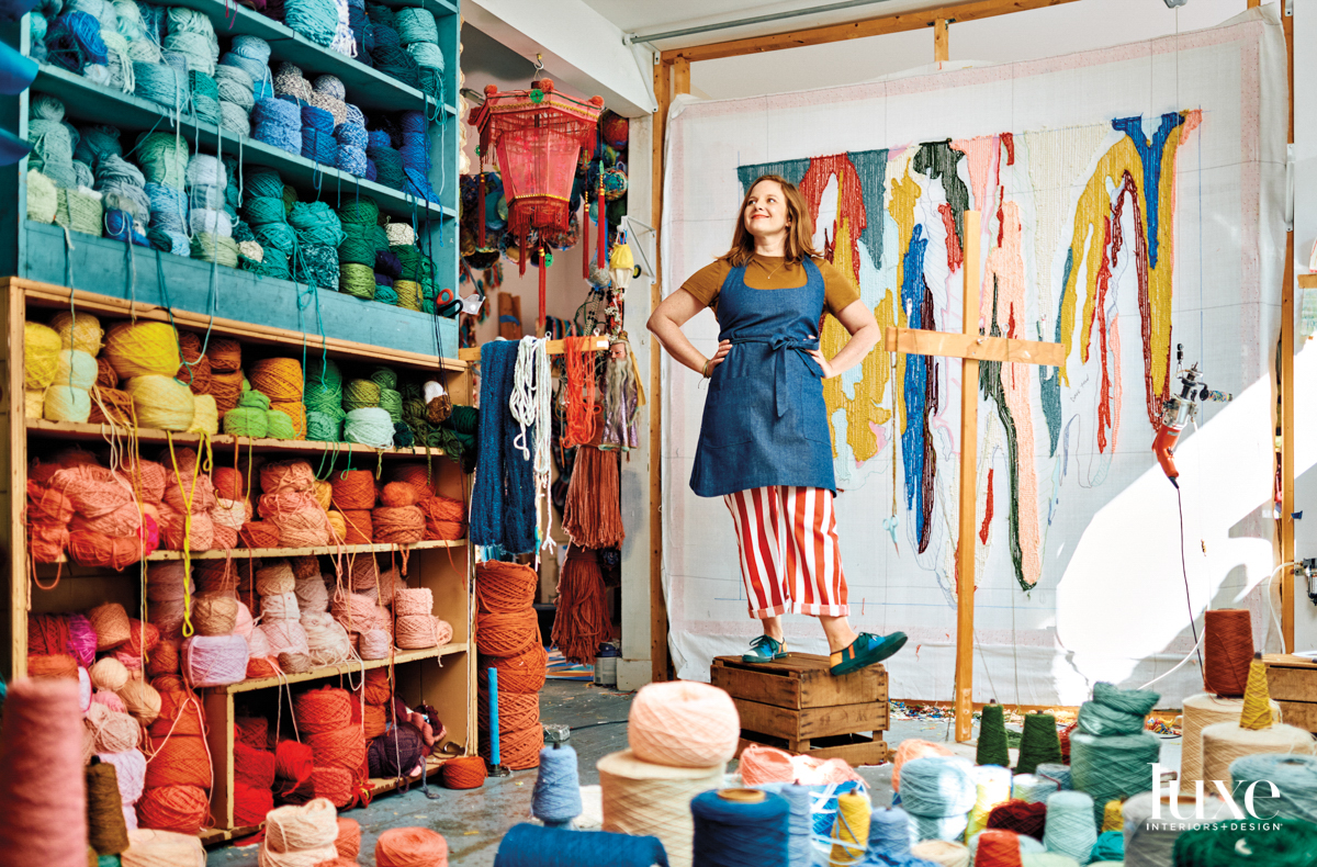 Woman wearing red striped pants and a denim apron standing in artist studio surrounded by hundreds of spools of colorful yarn