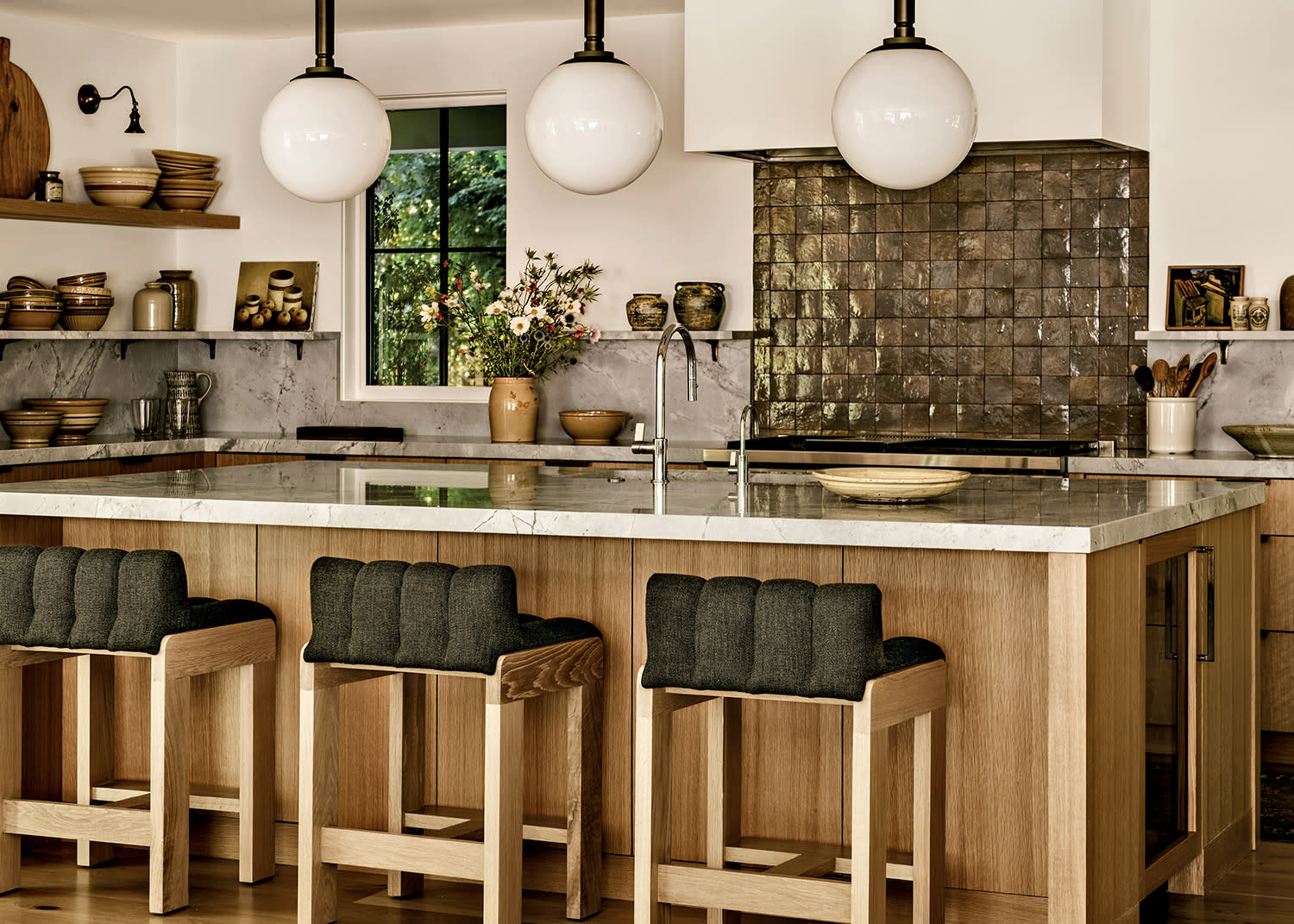 A cozy kitchen with a large marble island, wooden stools with dark cushions, globe pendant lights, and a textured tile backsplash. Shelves with rustic pottery add warmth.