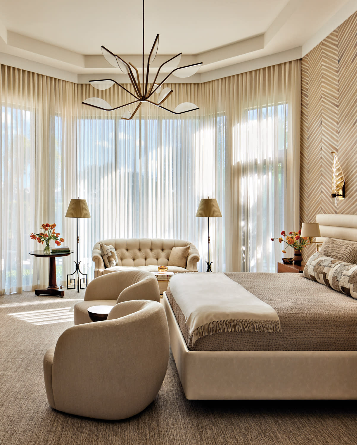 Spacious bedroom with beige tones, featuring a plush bed, cozy armchairs, tufted sofa, and tall windows with sheer curtains. Elegant, peaceful ambiance.