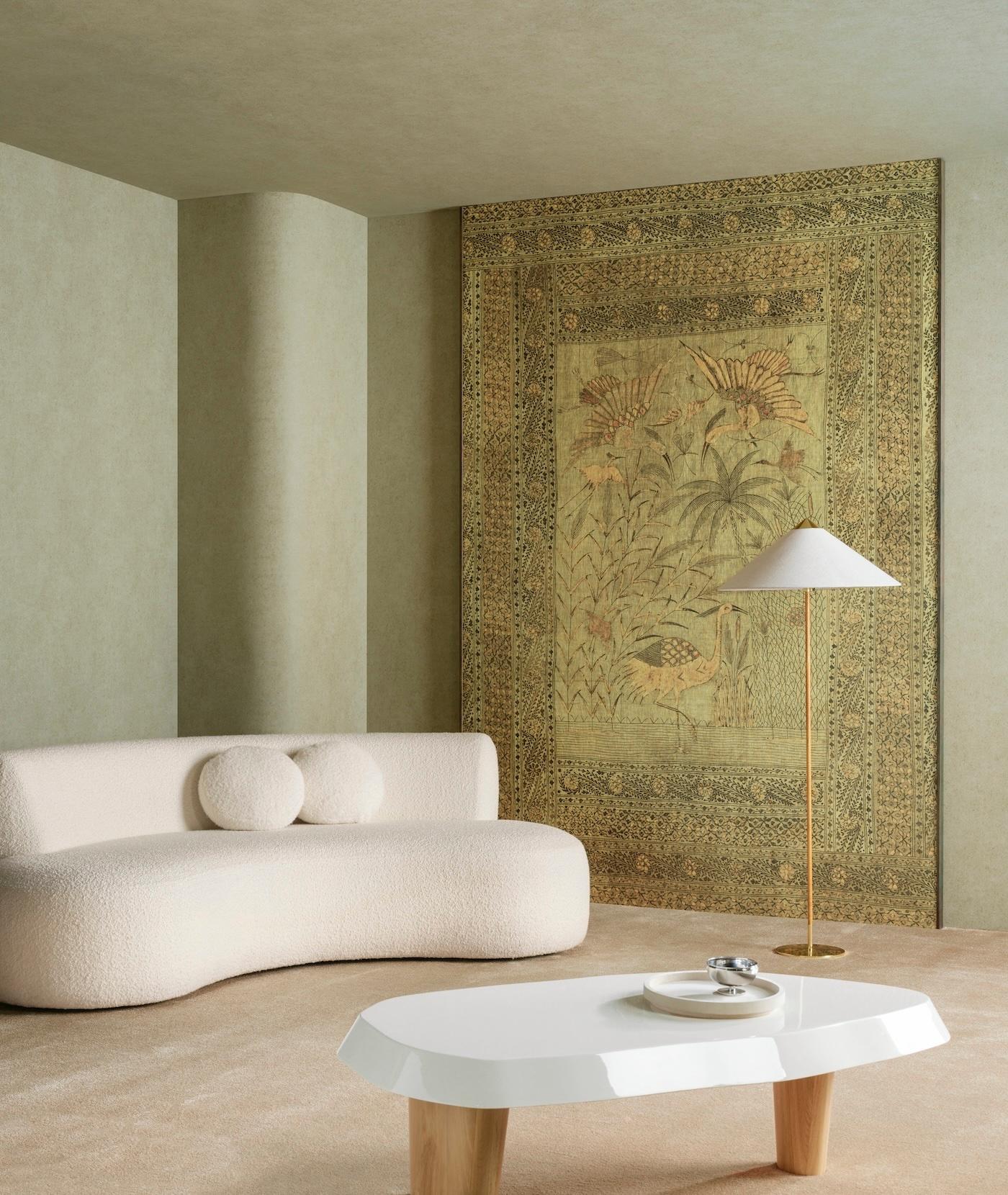 A beige modern living room featuring a curved sofa with round cushions, a sleek white coffee table, and a tall floor lamp. A large, ornate tapestry with floral and bird motifs is the focal point, creating a serene and elegant atmosphere.