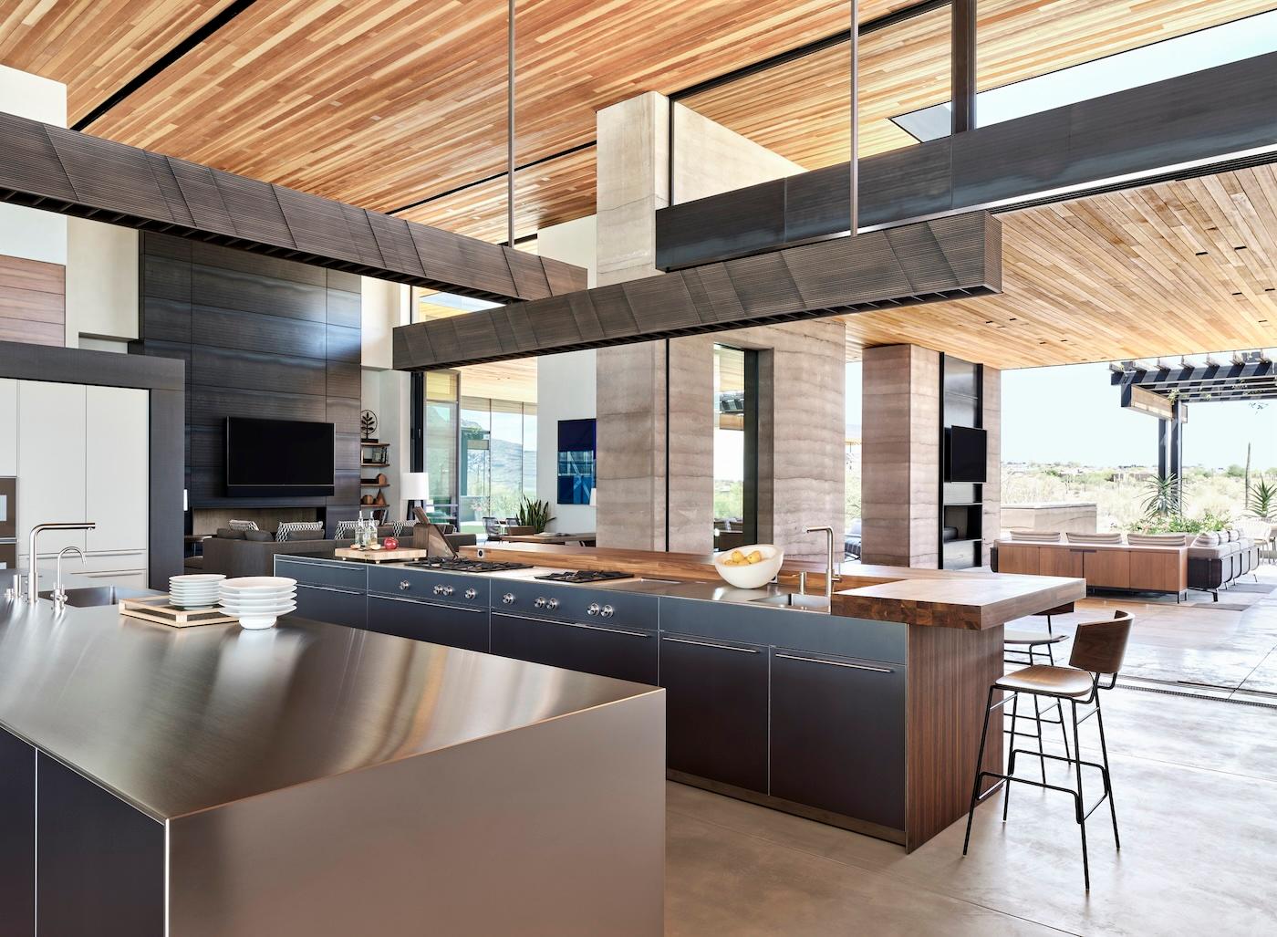 Spacious modern kitchen with sleek stainless steel countertops, wooden accents, bar stools, and large windows providing natural light and an airy feel.