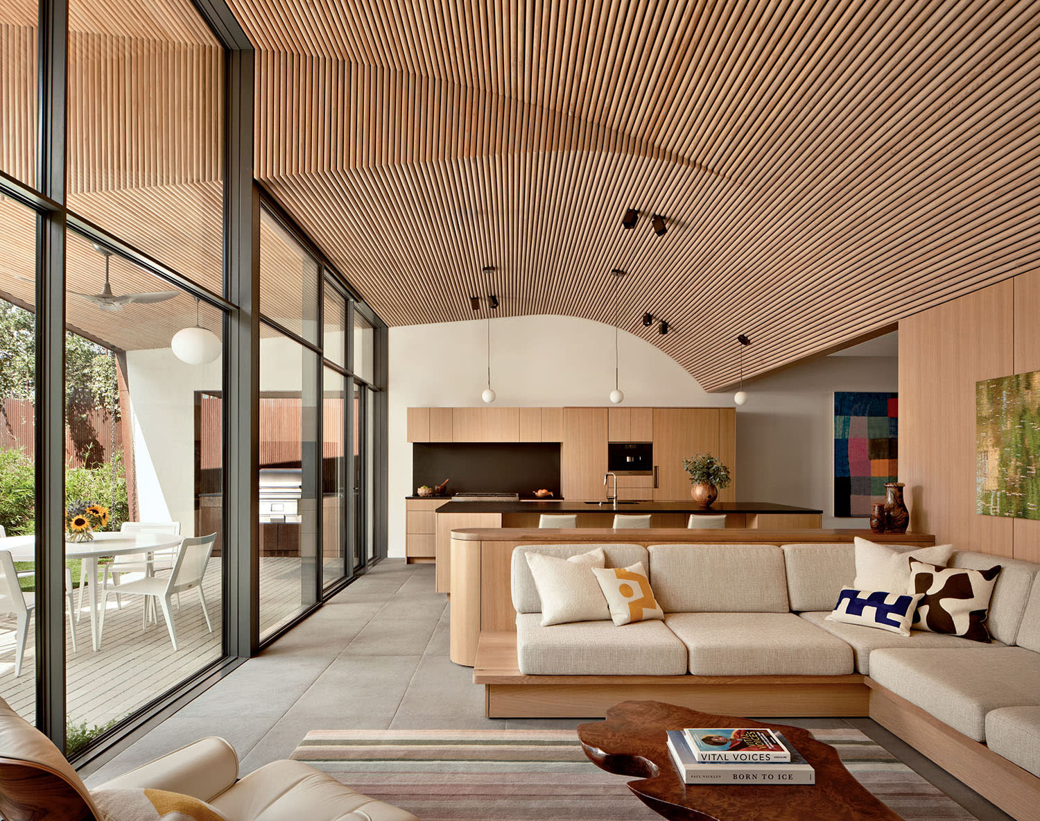 Modern open-plan living space with wooden slatted ceiling, beige sofa, colorful cushions, sleek kitchen island, and large windows opening to a patio.