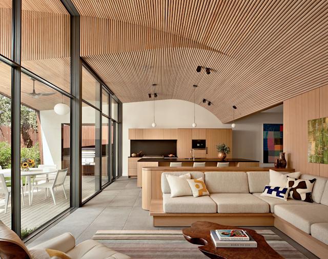 Modern open-plan living space with wooden slatted ceiling, beige sofa, colorful cushions, sleek kitchen island, and large windows opening to a patio.
