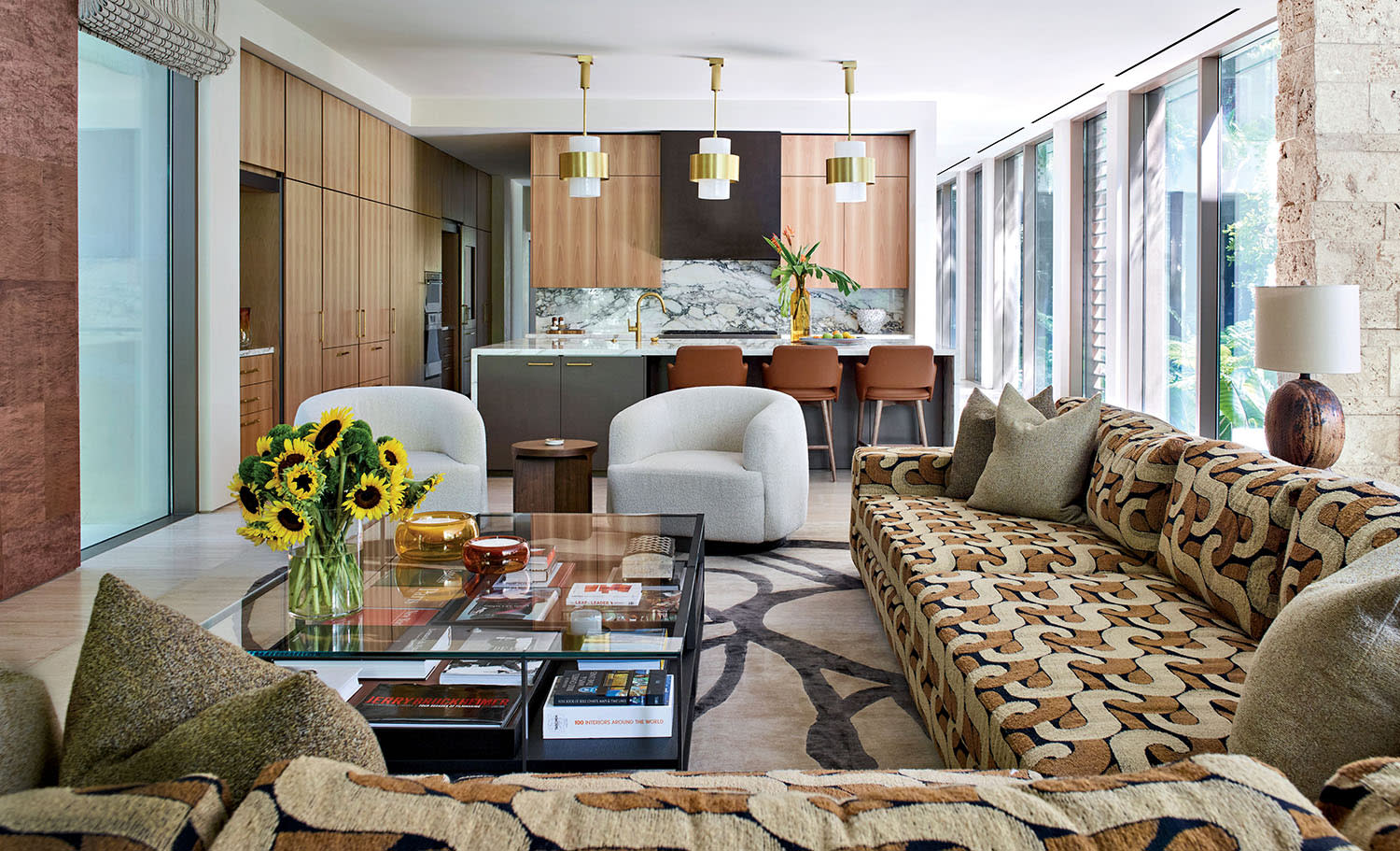 Spacious modern living room with a patterned sofa, glass coffee table with sunflowers, and an open kitchen with wood cabinets and pendant lights.