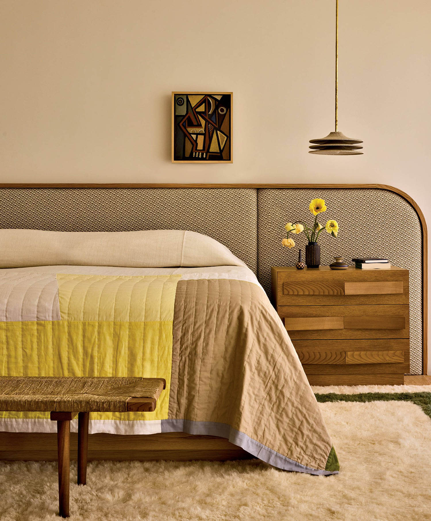 A bed sits against a fabric headboard that curves past a wood nightstand, with a bench at its foot.