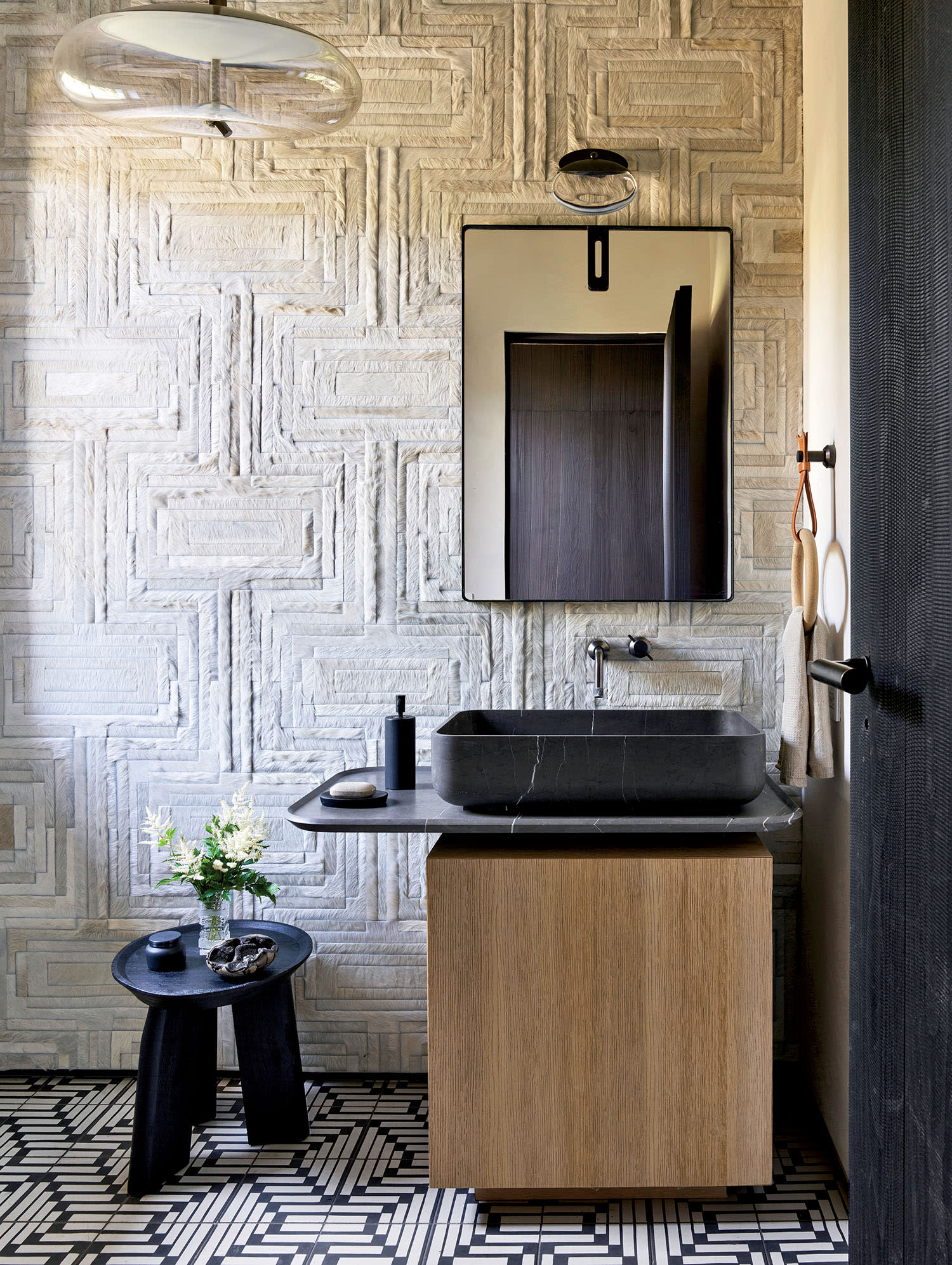 Modern bathroom with textured beige walls, black patterned tile floor, wooden vanity with black basin, round mirror, and a vase of flowers on a small black table.