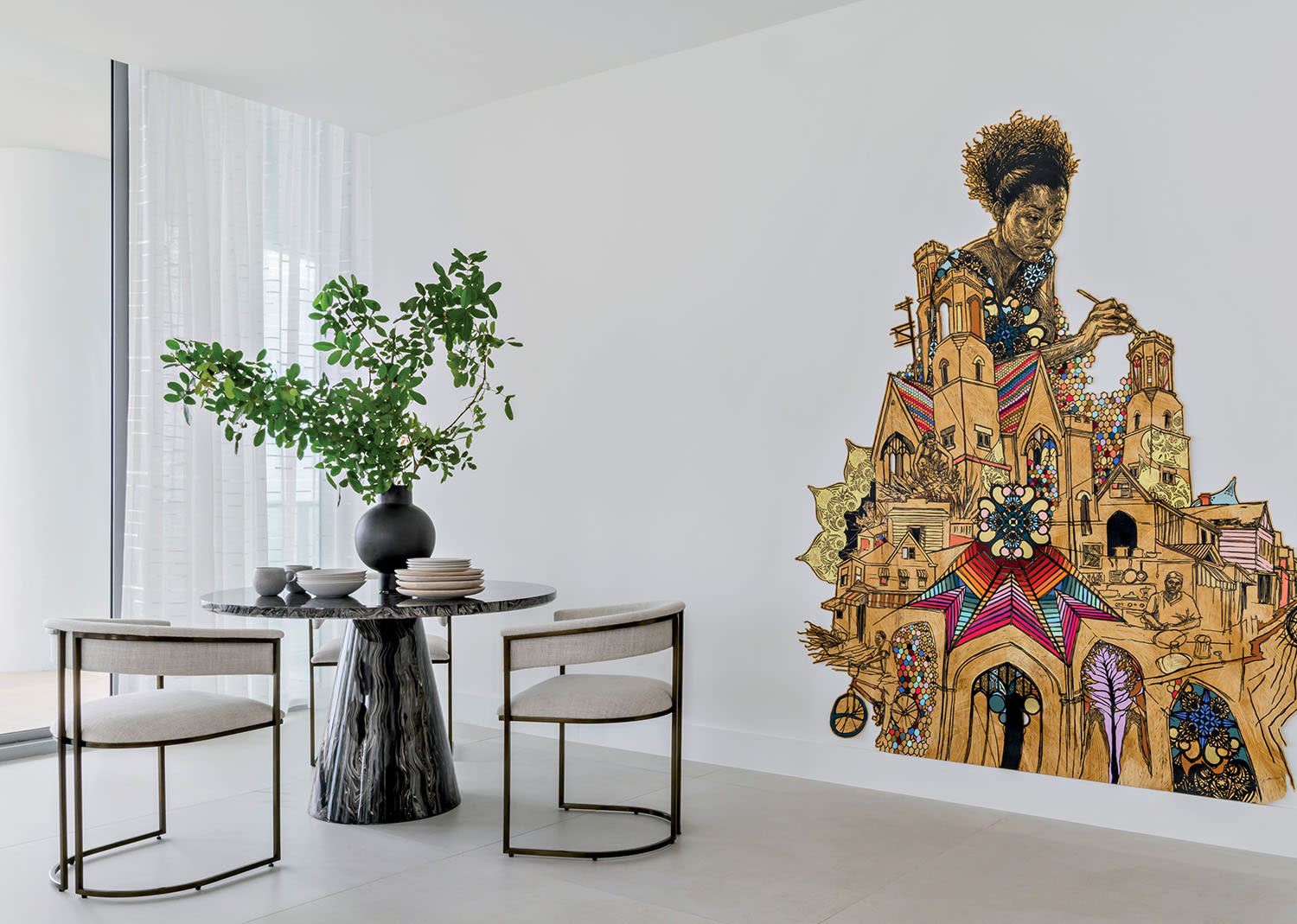 A modern dining area with a round table, two chairs, and a vase with green branches on the table. The wall features a colorful mural of an intricate cityscape with a large, expressive face at the top