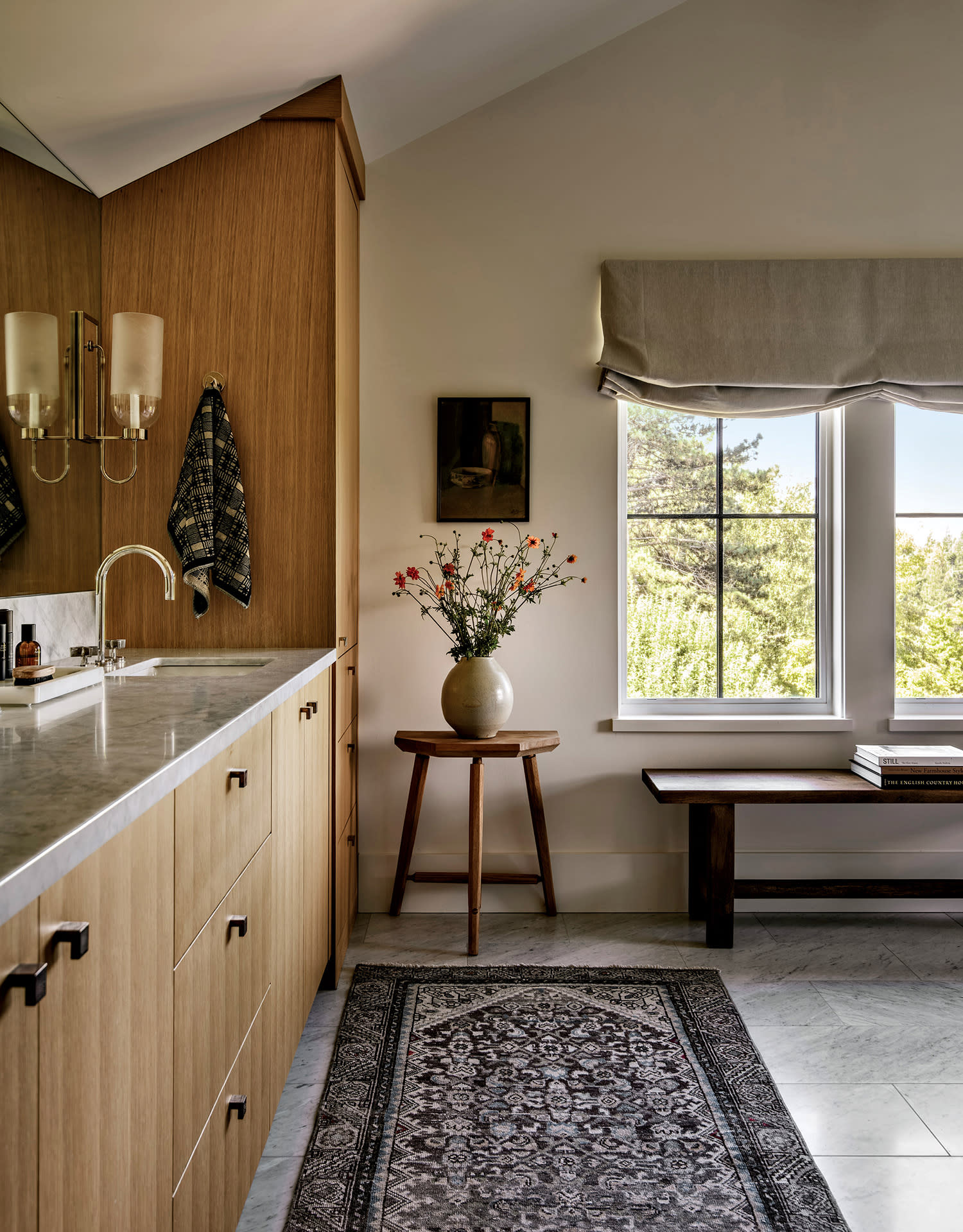 A serene bathroom with a wooden vanity, marble countertop, and wall-mounted lights. A potted flower vase sits near a window with a nature view.