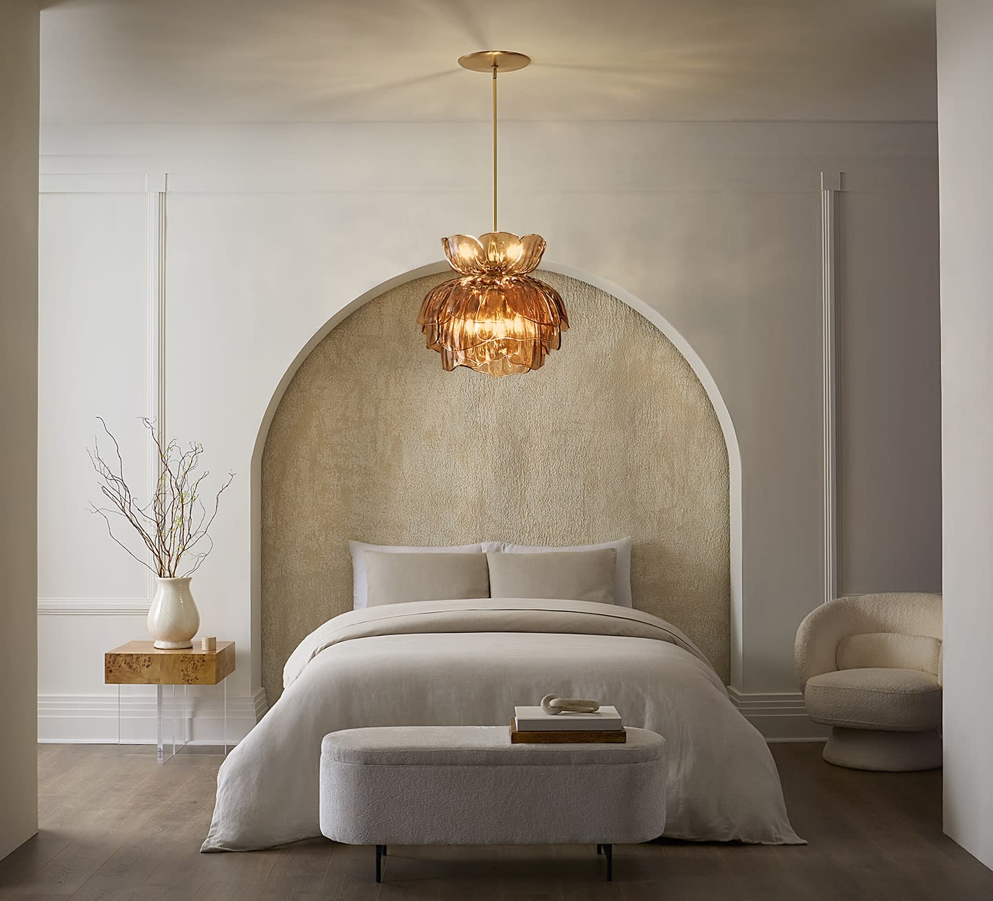 Elegant bedroom with a cozy bed under a textured archway. A warm, golden chandelier hangs above. Minimalist decor includes a side table with a vase.
