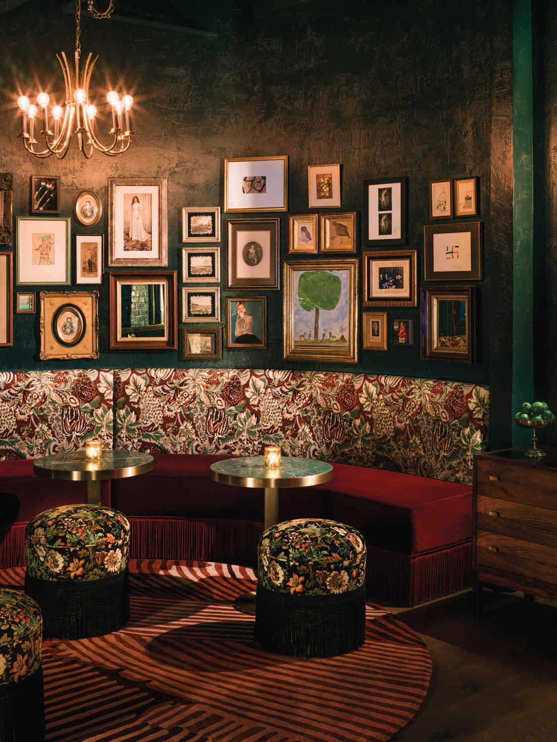 Curved banquette with red floral upholstery beneath a gallery wall and paired with cocktail tables