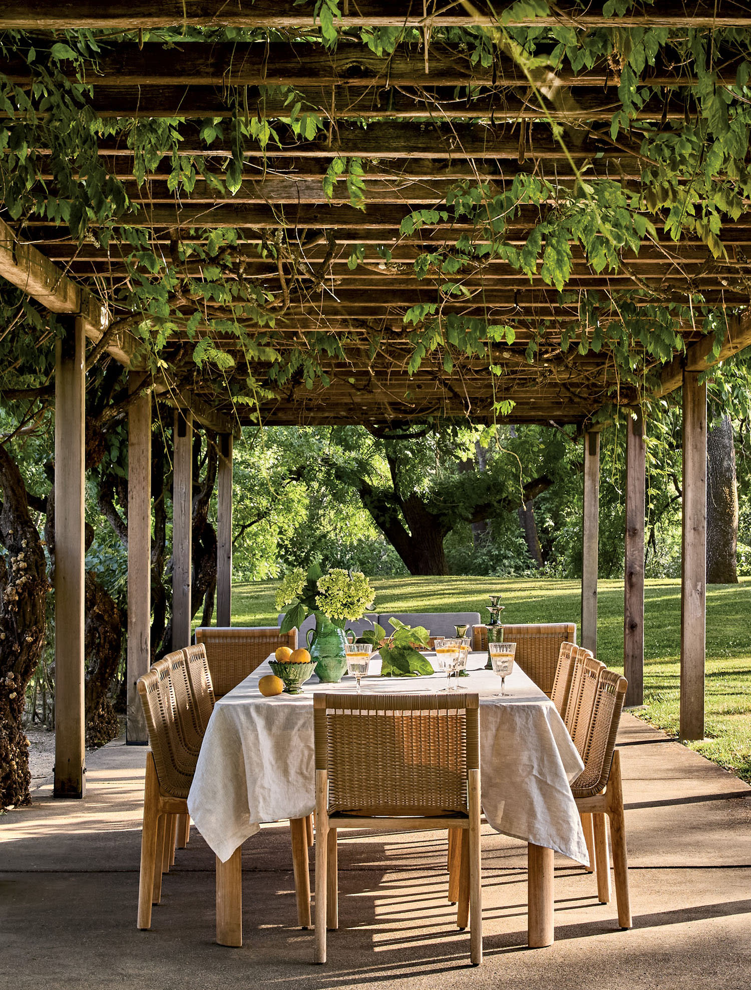 Outdoor dining under a wooden pergola adorned with lush vines, featuring a table with a white cloth, flowers, and lemons, evoking a serene, welcoming atmosphere.