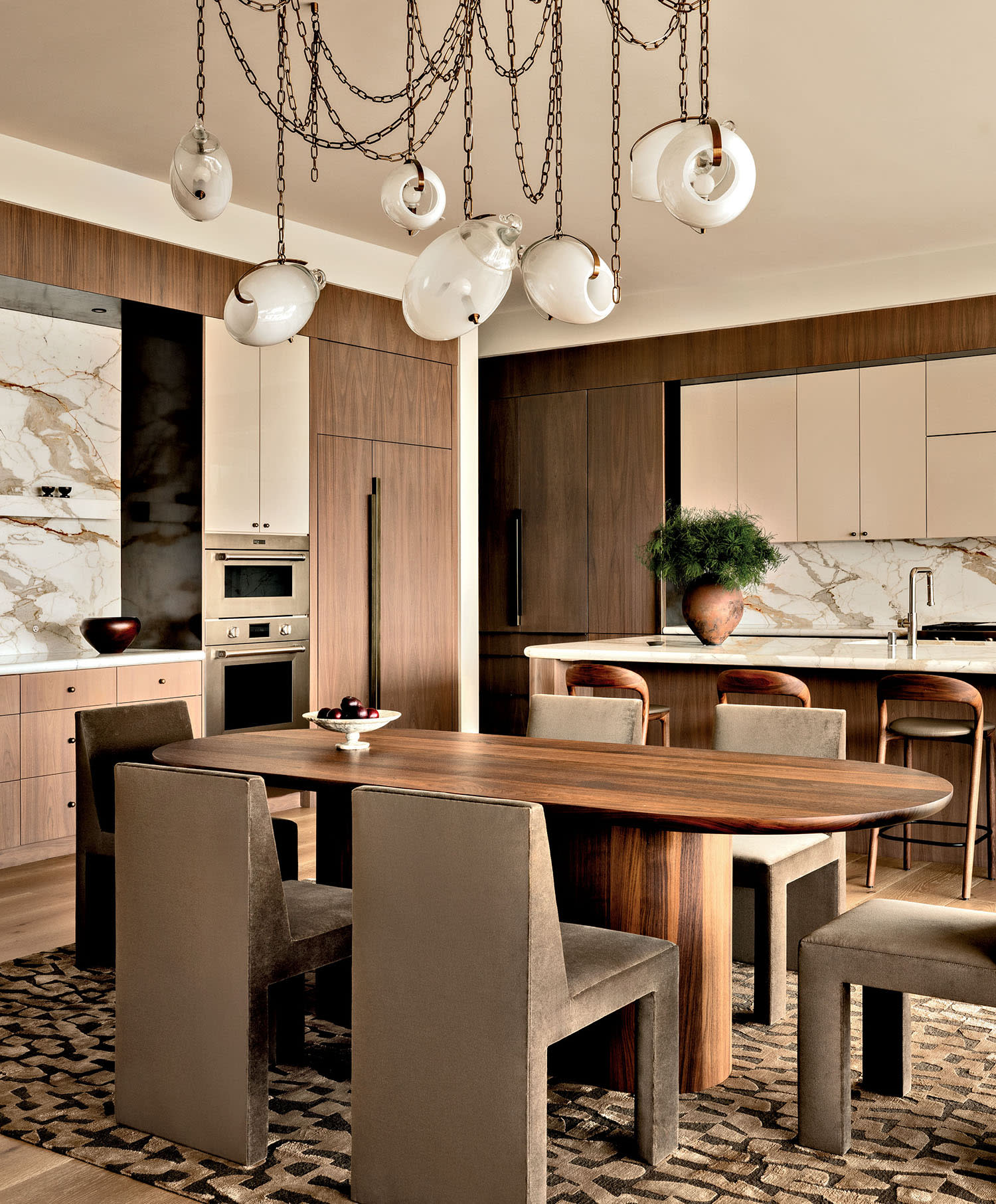 An abstract chandelier with chains hangs above a wood dining table set with six chairs near the kitchen.