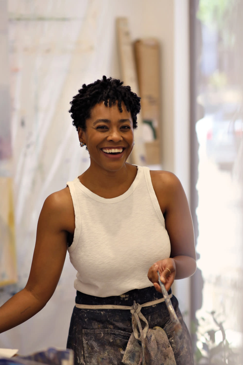 Arielle Austin laughing while holding a paint brush in a white tank top and apron.