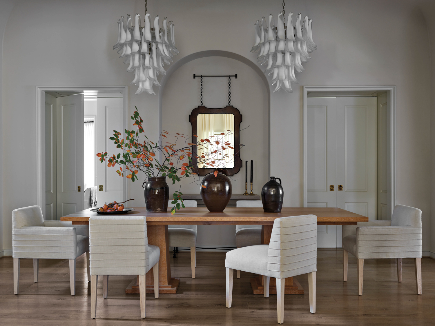 White dining room with wooden table, upholstered chairs and double chandeliers by Anna Booth