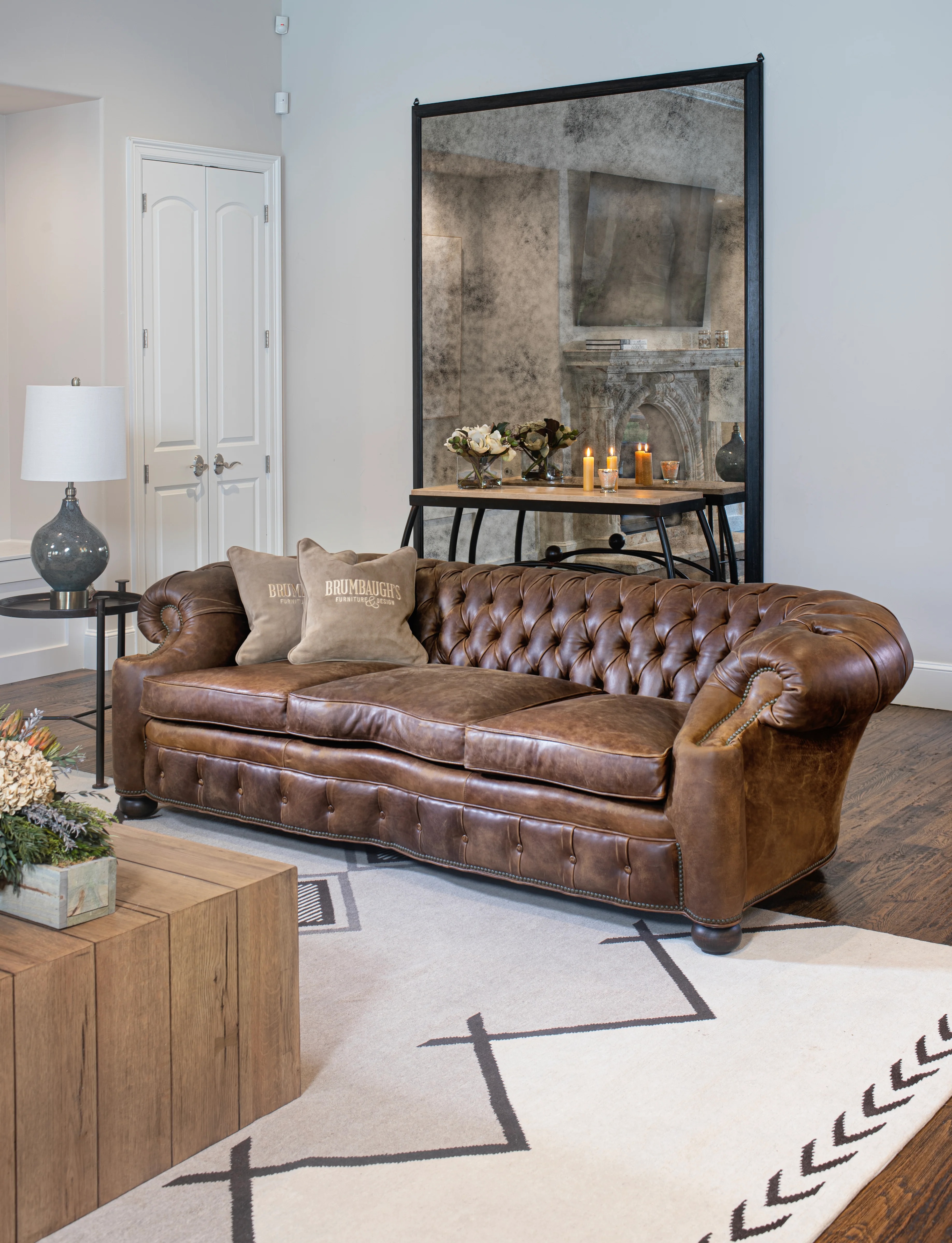 Luxurious living room with a tufted brown leather sofa, soft cushions, a large mirror reflecting a fireplace, and a rustic wooden coffee table.