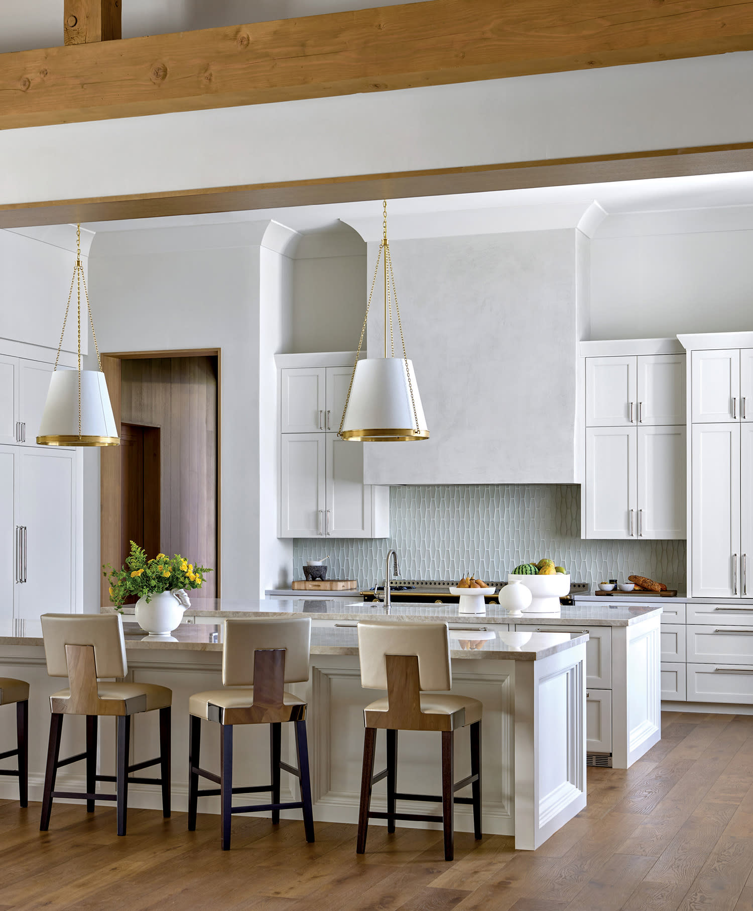 Spacious modern kitchen with white cabinets, wood beams, and a large island. Pendant lights hang above the island, with stools and a wooden floor.