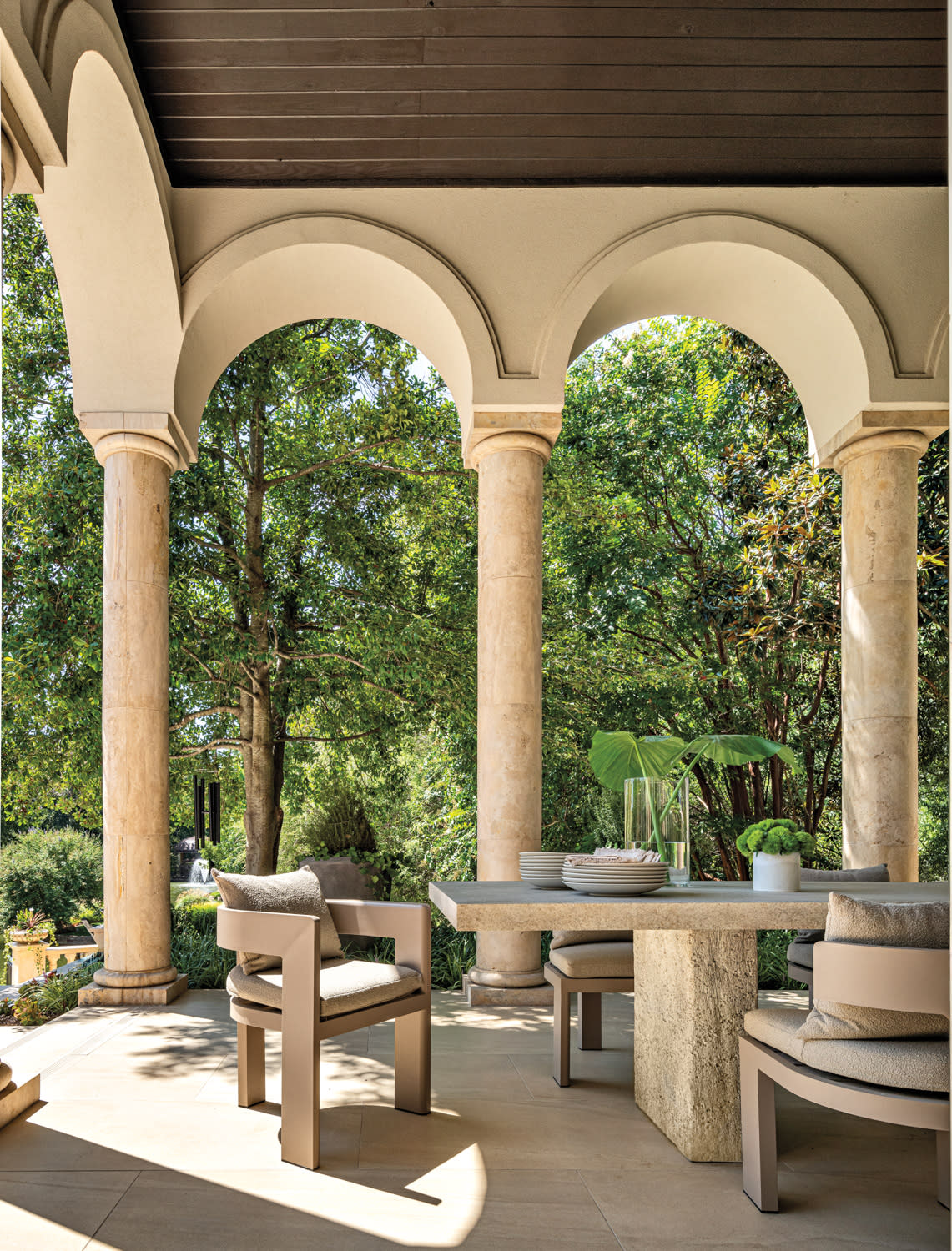 Elegant patio with beige stone pillars and arches, earthy-toned furniture, and lush greenery in the background, conveying a serene, classic ambiance.