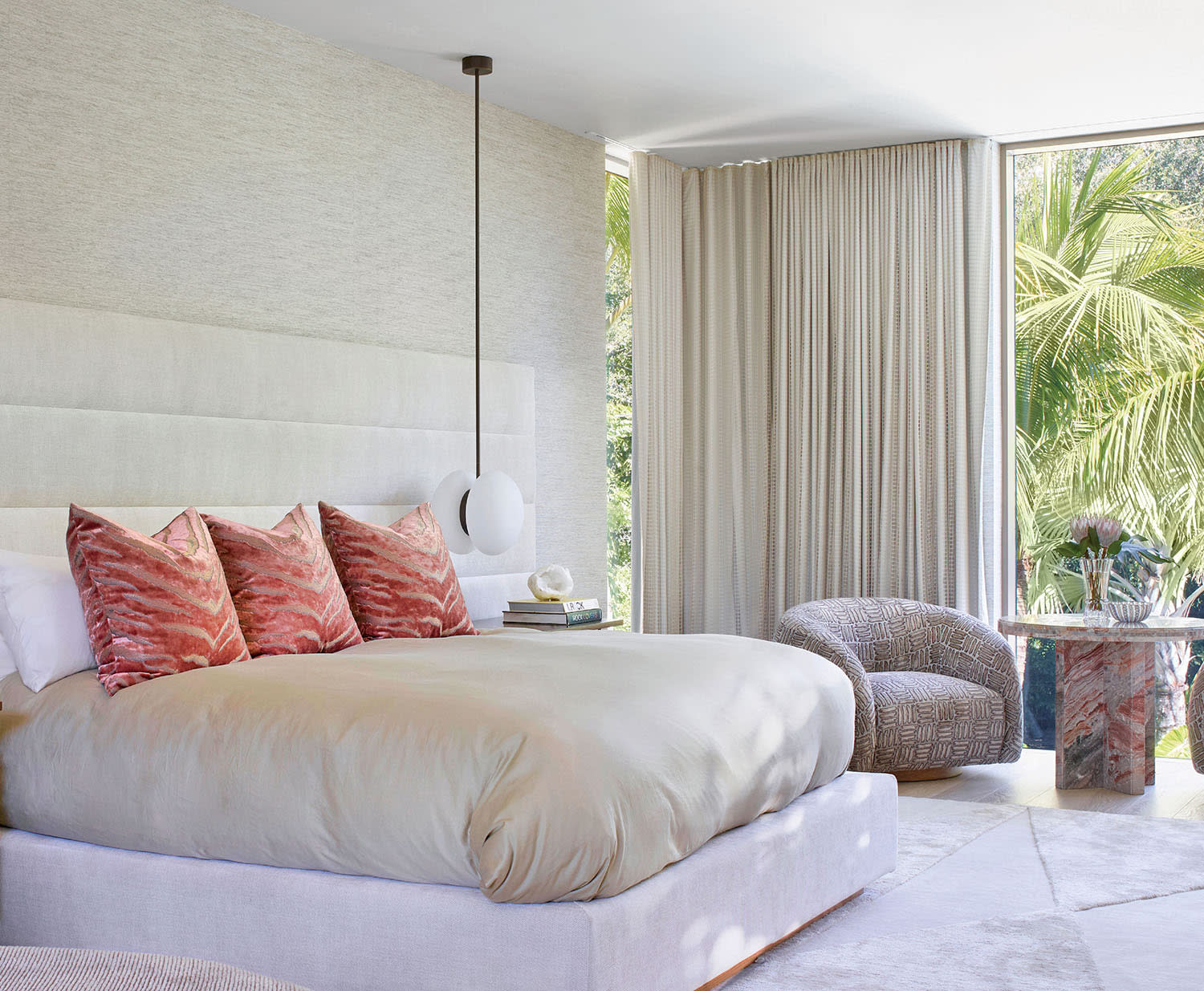 Modern bedroom with a large bed featuring beige and red pillows, elegant pendant lights, and cozy armchairs. Floor-to-ceiling windows offer a garden view.
